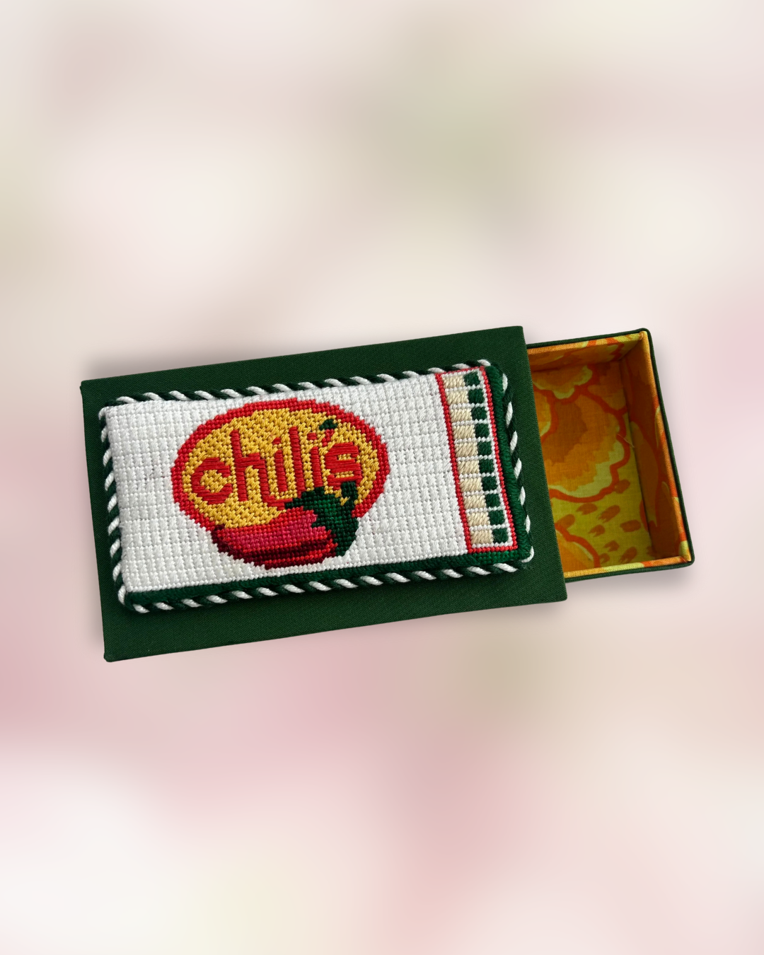 A matchbox-style decorative box with an embroidered chili pepper and the word 'chili' on the lid, partially open revealing a orange floral pattern interior.