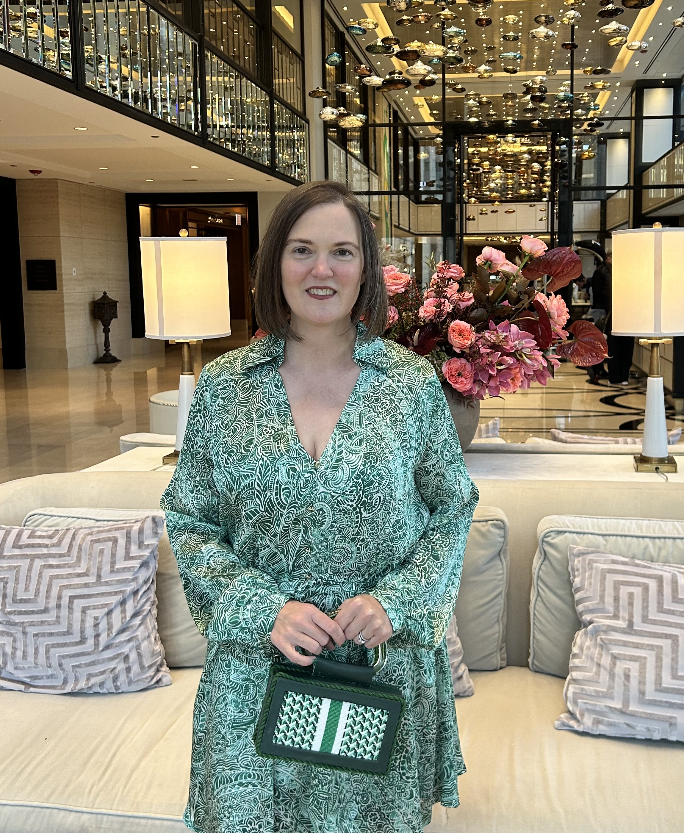 A woman with shoulder-length brown hair wearing a green patterned dress, holding a green and white handbag, standing in a hotel lobby with a beige sofa, pink flower arrangement, and decorative ceiling lights.