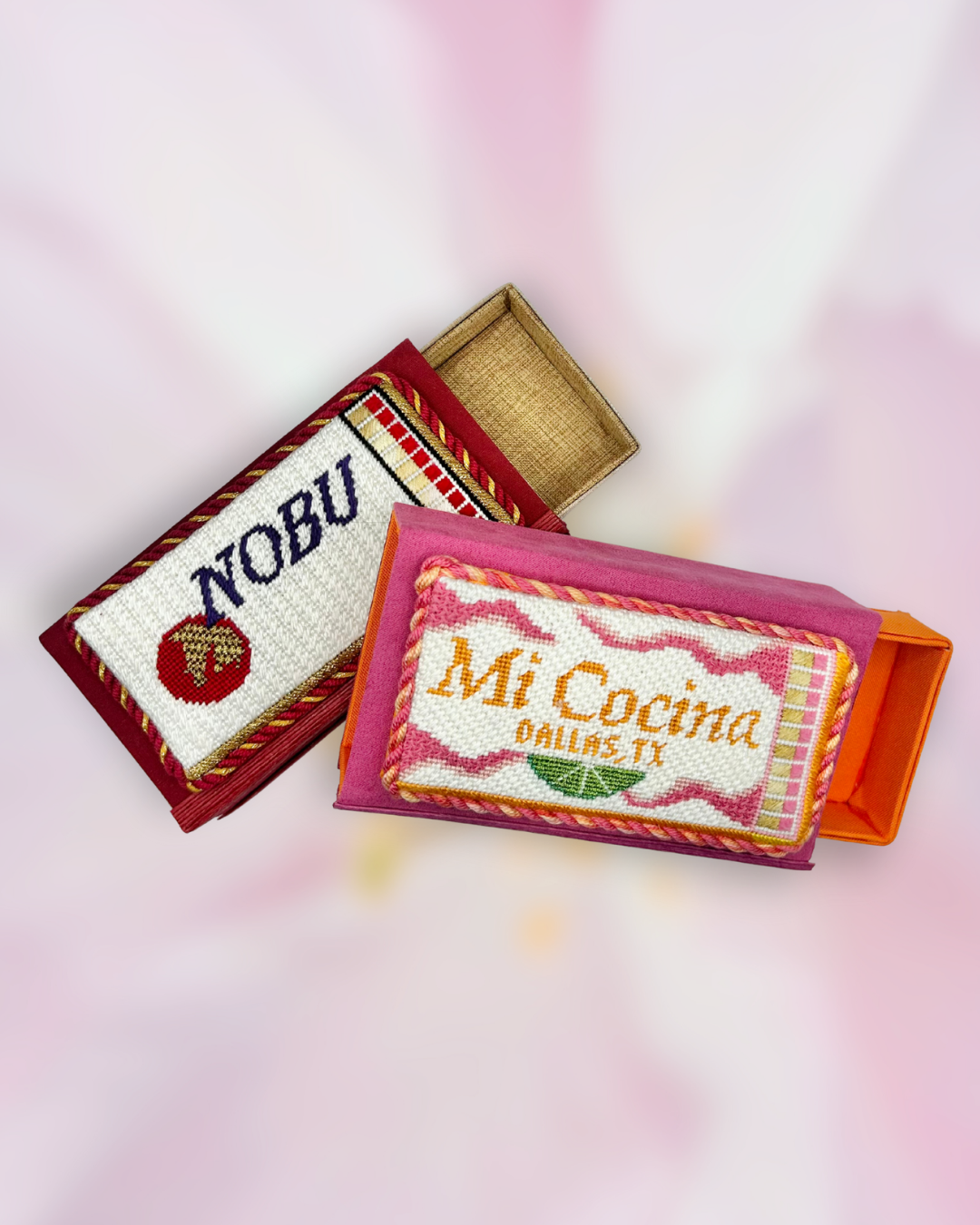 Two embroidered needlepoint matchbox cases, one with a 'NOBU' logo and the other with 'Mi Cocina Dallas TX', on a blurred pink background.