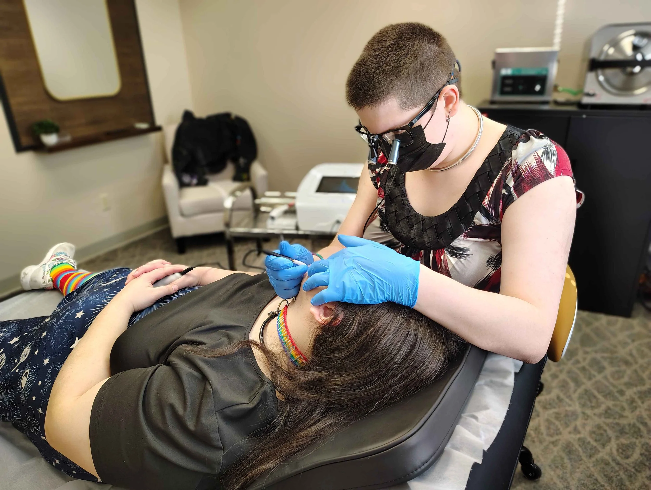 Electrologist wearing gloves, mask, and magnifying glasses, probe in hand, tattooing a young woman lying on a table with a rainbow-colored choker, in an electrolysis clinic.