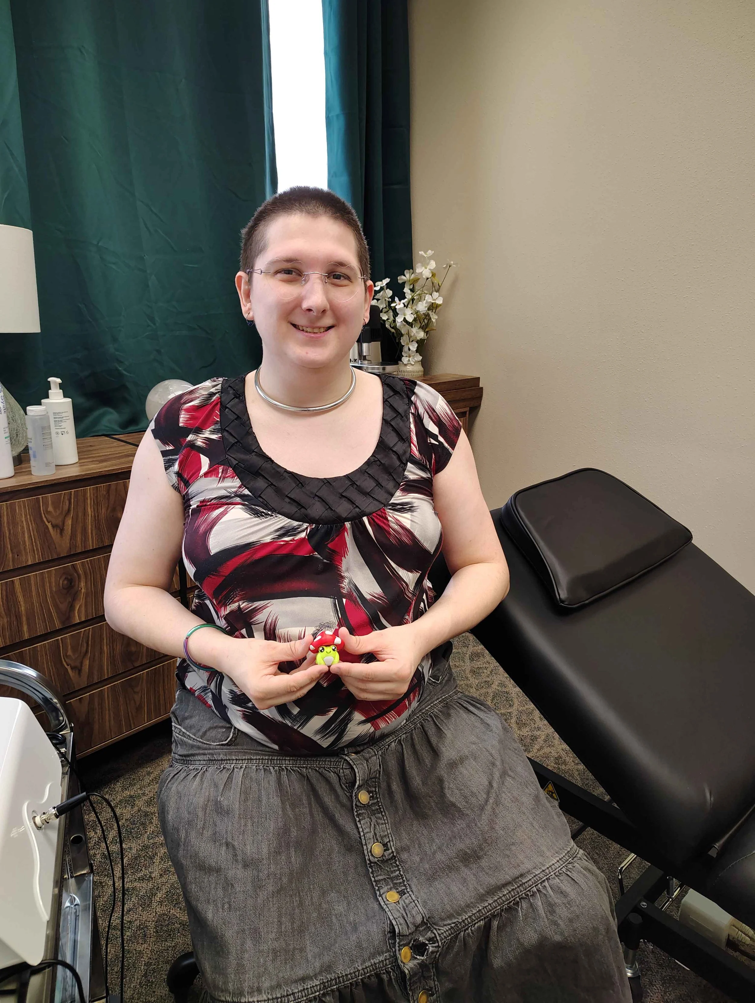 A woman sitting in a medical office, smiling, holding a small frog clicker, with a black treatment table next to her, and a wooden cabinet with bottles in the background.
