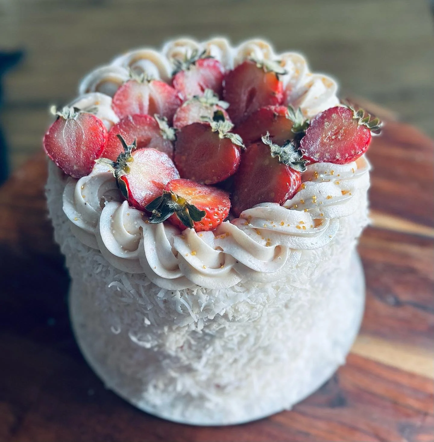 A dessert topped with halved strawberries and decorated with swirls of white frosting, golden sprinkles, and shredded coconut on a wooden surface.