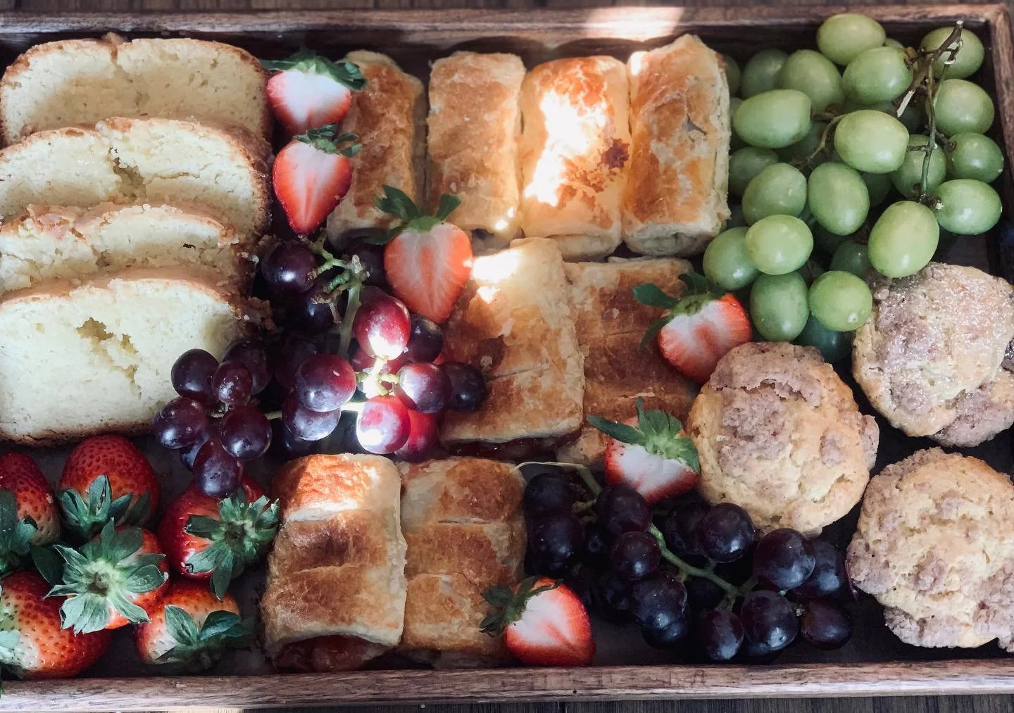 Assorted pastries, strawberries, green grapes, and purple grapes in a wooden tray.