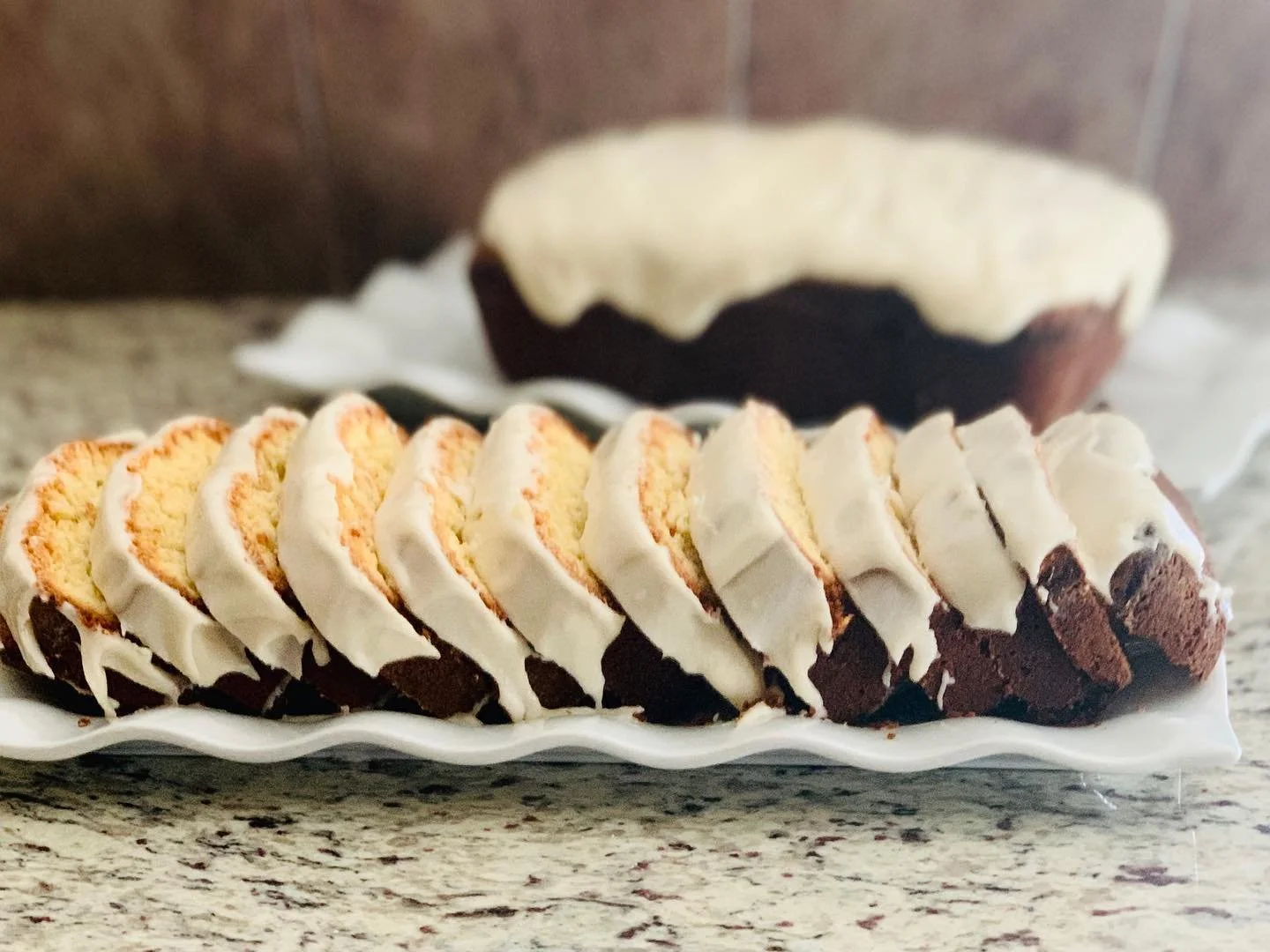 A plate of sliced cake with white frosting and a chocolate base, with a larger whole cake with similar frosting and chocolate layers in the background on a marble countertop.