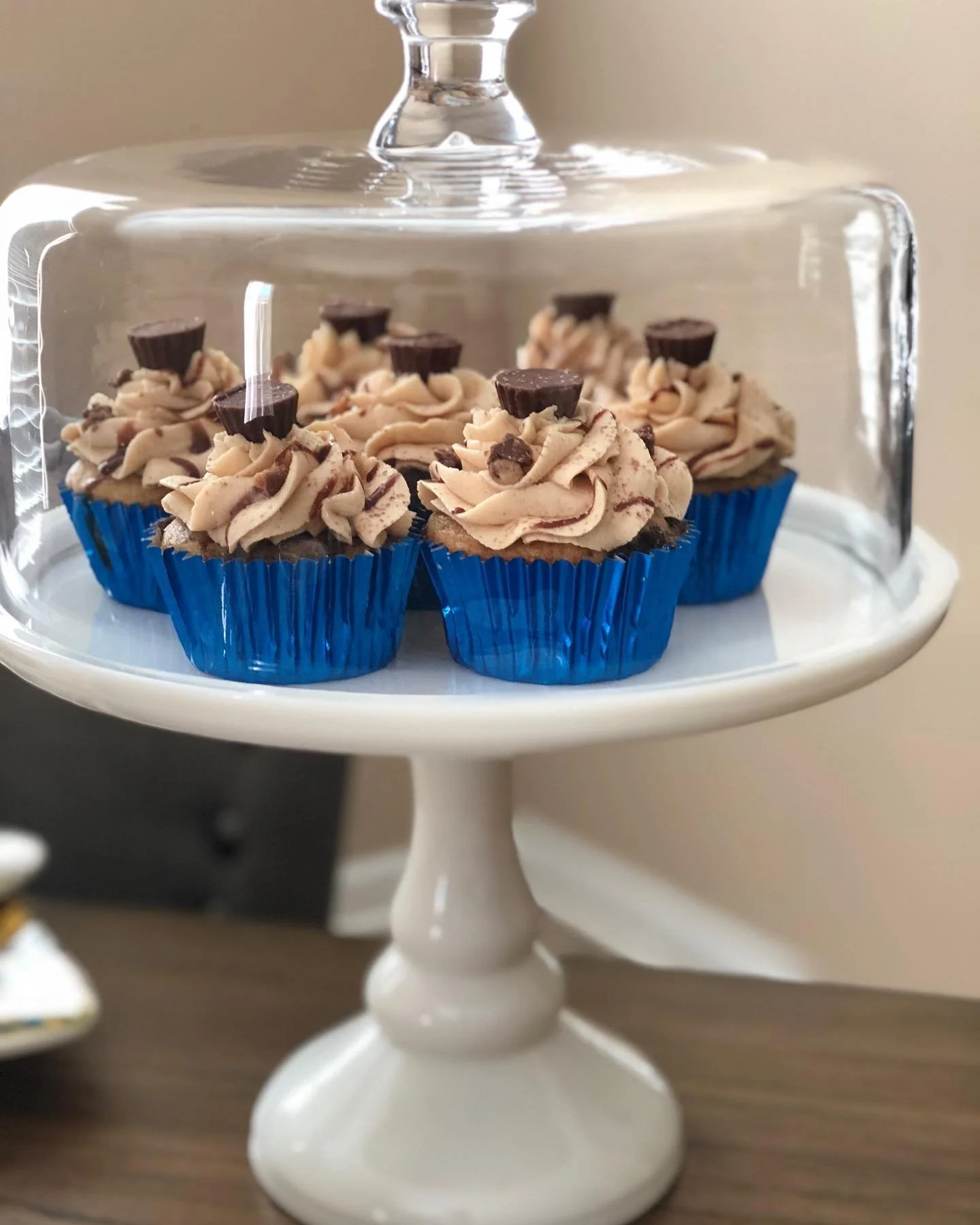 Chocolate cupcakes with swirled frosting and small chocolate pieces on a white cake stand with a glass cover.
