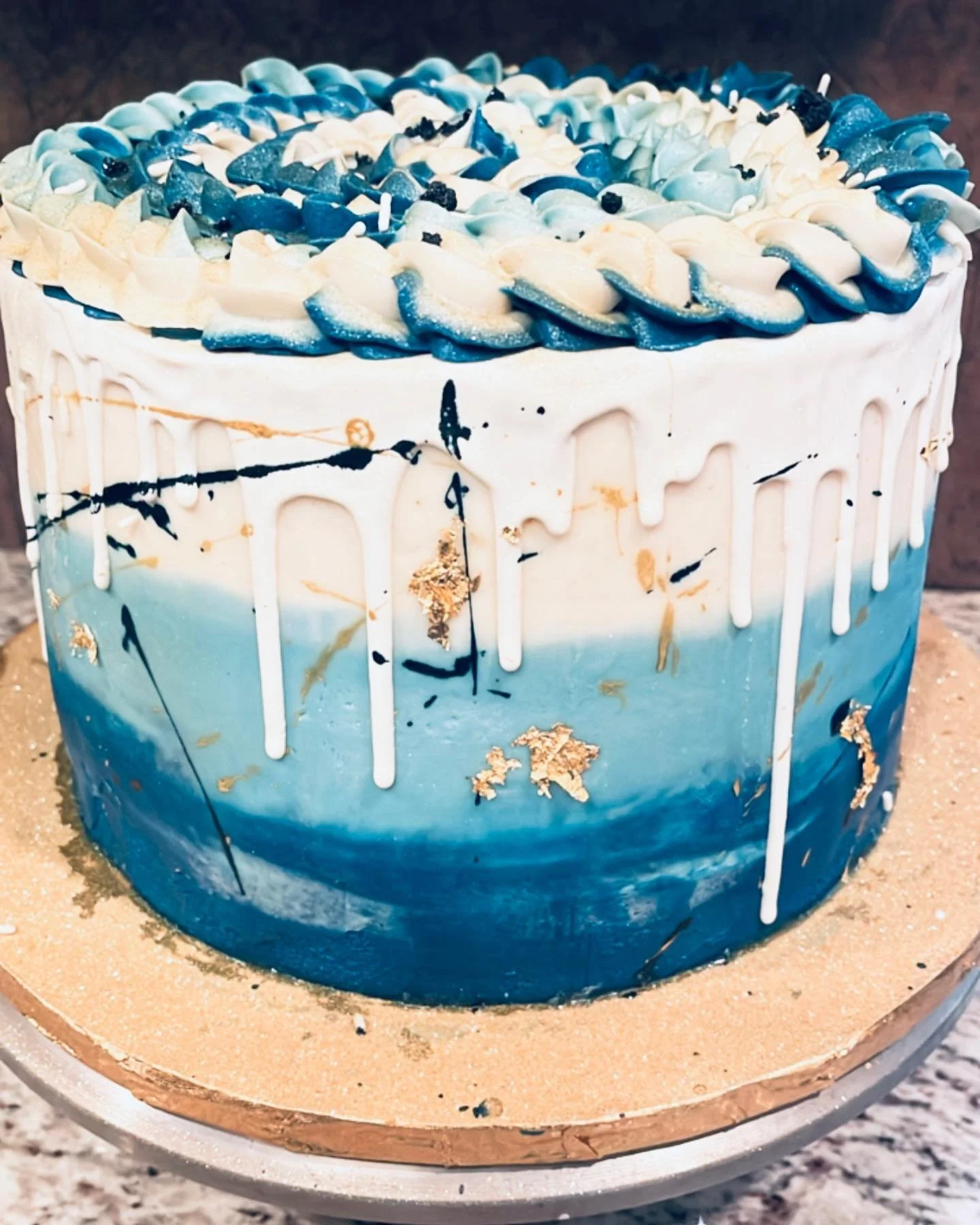A round cake with blue and white ombre icing, gold leaf decorations, and blue and white buttercream piping on top, with paint splatter-style black and blue accents.