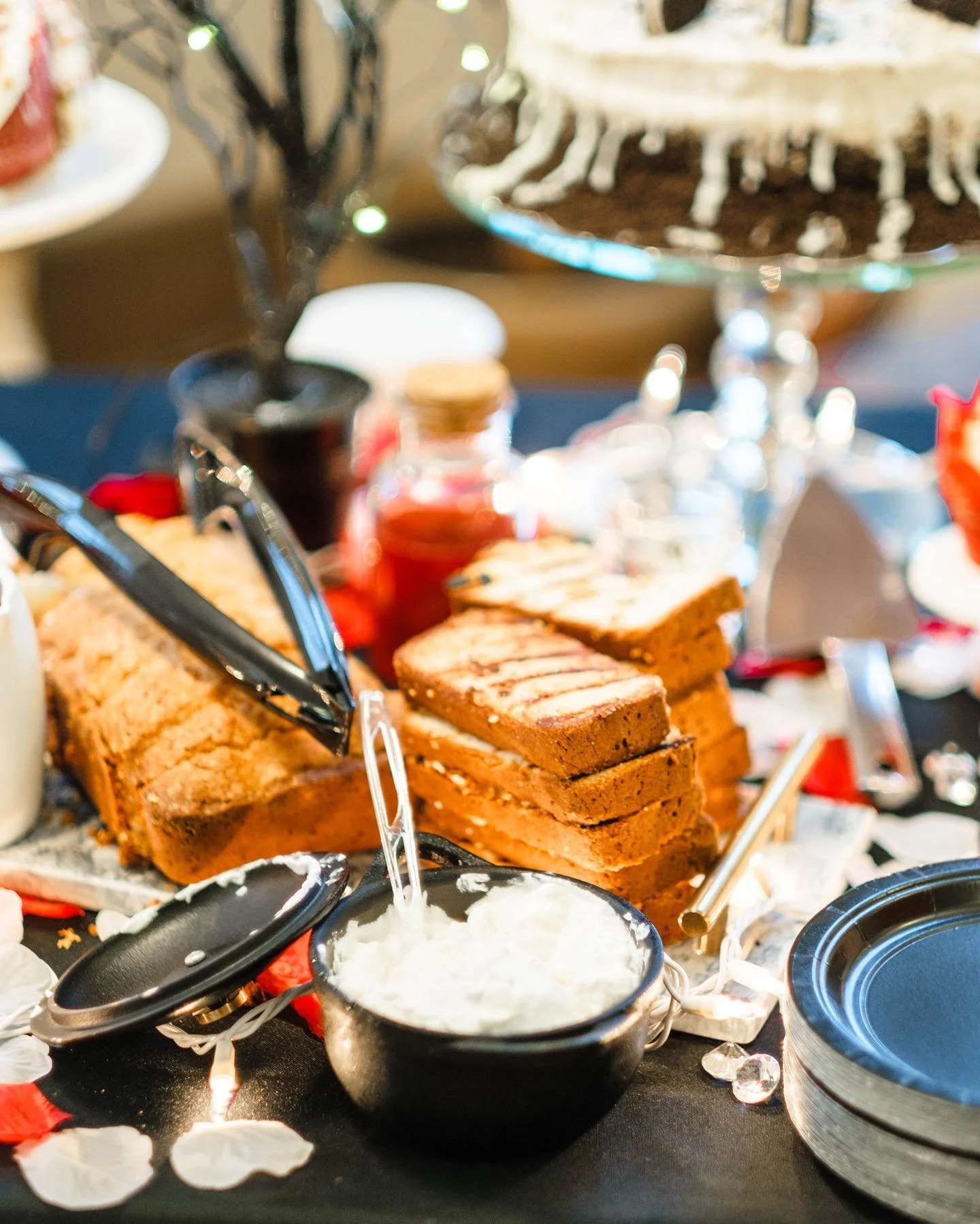 Assorted cakes and desserts on a table, including a sliced loaf cake, stacked blondies or brownies, a small bowl of whipped cream, and decorative rose petals.
