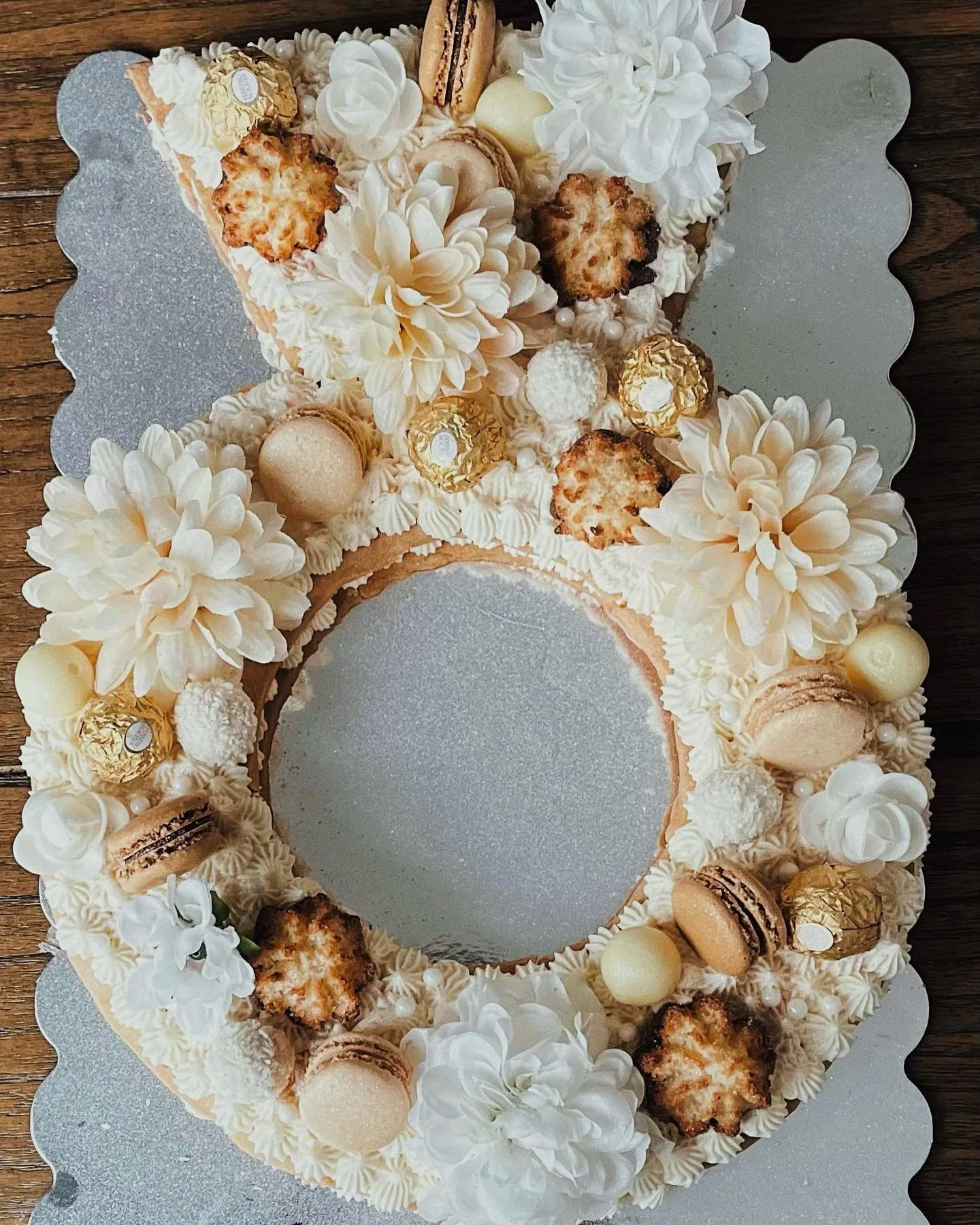 Cake decorated with white flowers, macarons, chocolates, and confections, with a hollow center on a silver board.