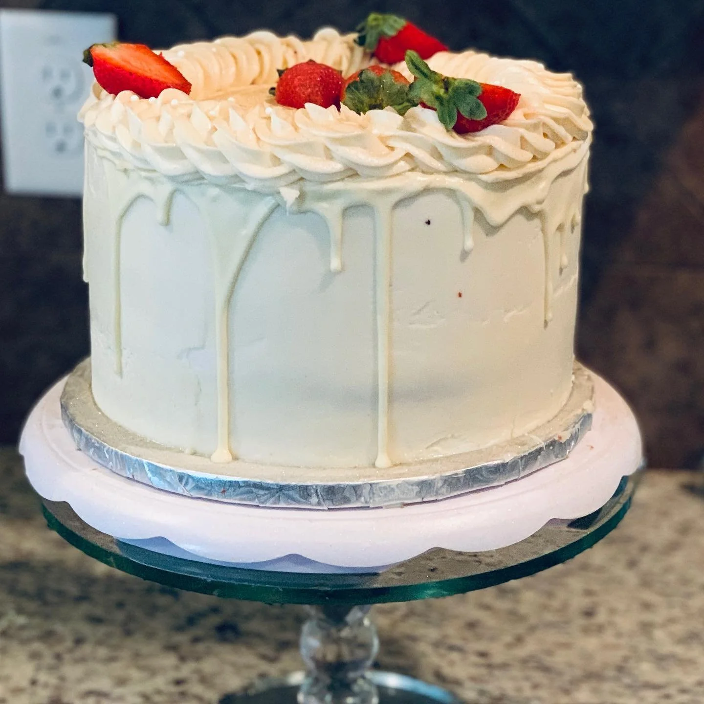 White frosted cake with strawberries and greenery on top, decorated with a white drip effect, placed on a glass cake stand.