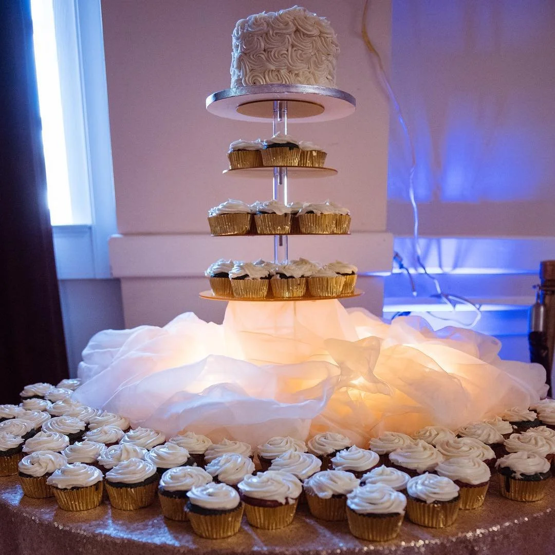 A tiered cupcake stand with a white, ruffled cake on top, surrounded by cupcakes with white frosting, beneath a glowing cloth and decorative lights inside a room with a window and blue ambient lighting.