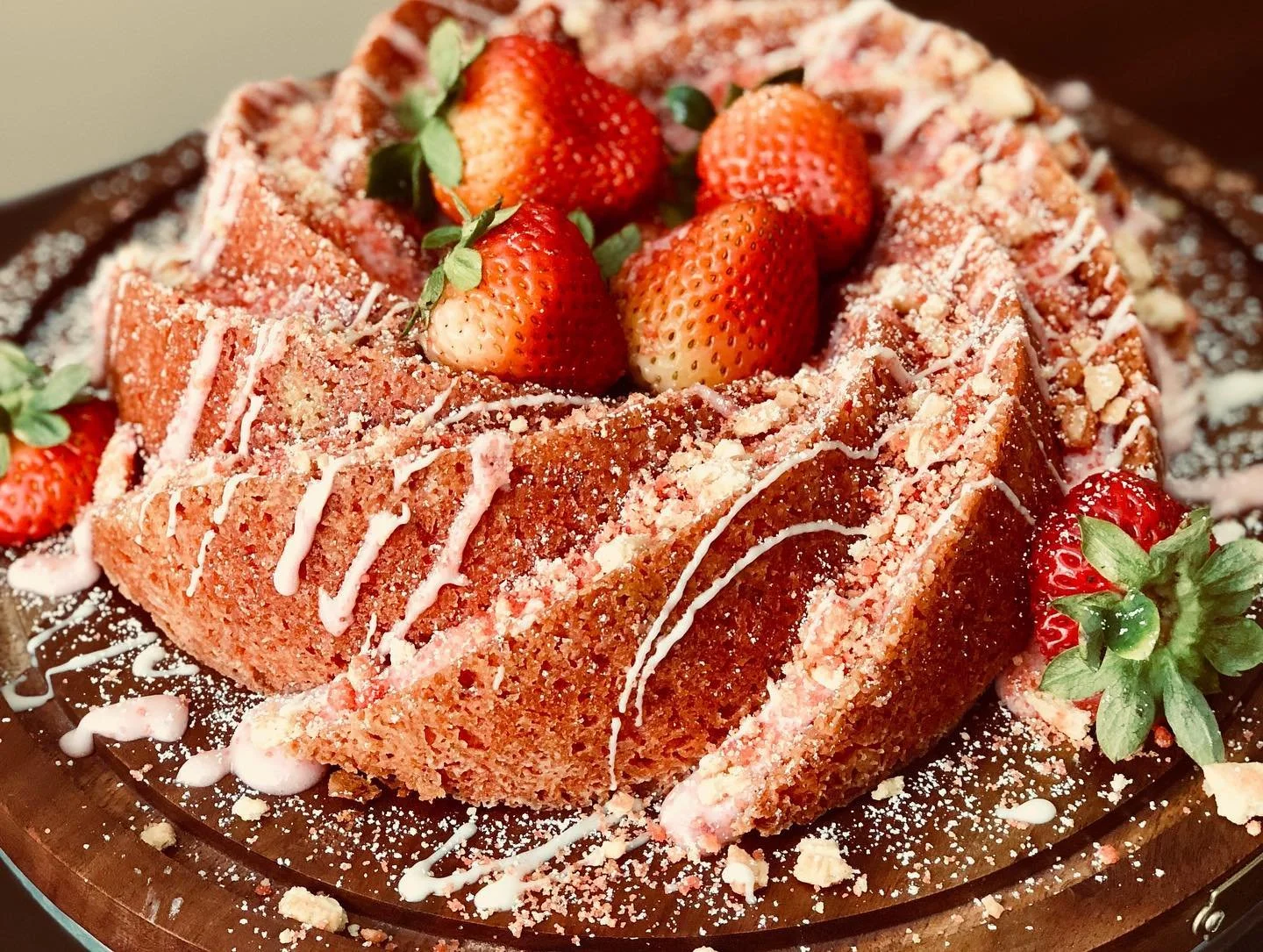 A square slice of strawberry cake topped with fresh strawberries, white chocolate drizzle, and sprinkled with crushed nuts or crumbs, served on a wooden board.