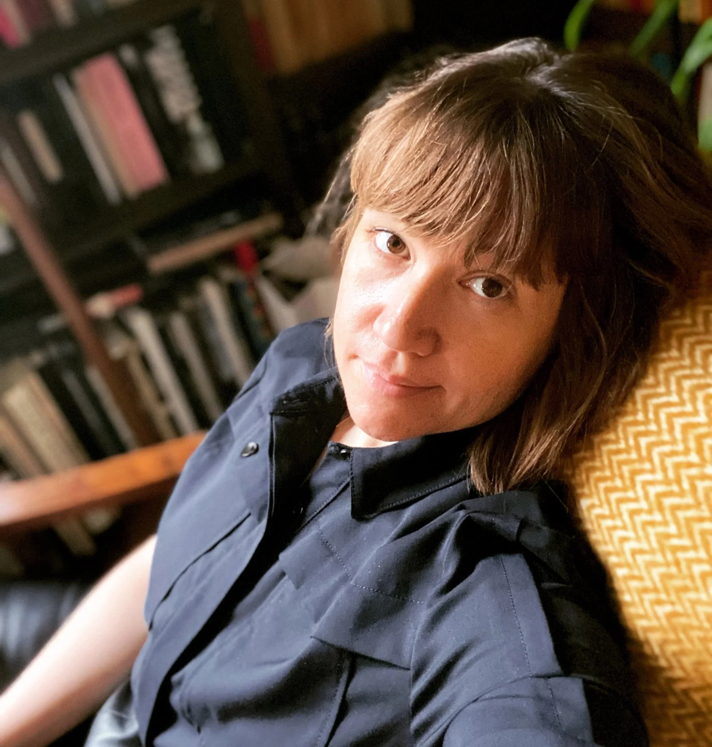 A person with brown hair and light skin, sitting against a patterned yellow cushion with a bookshelf in the background.