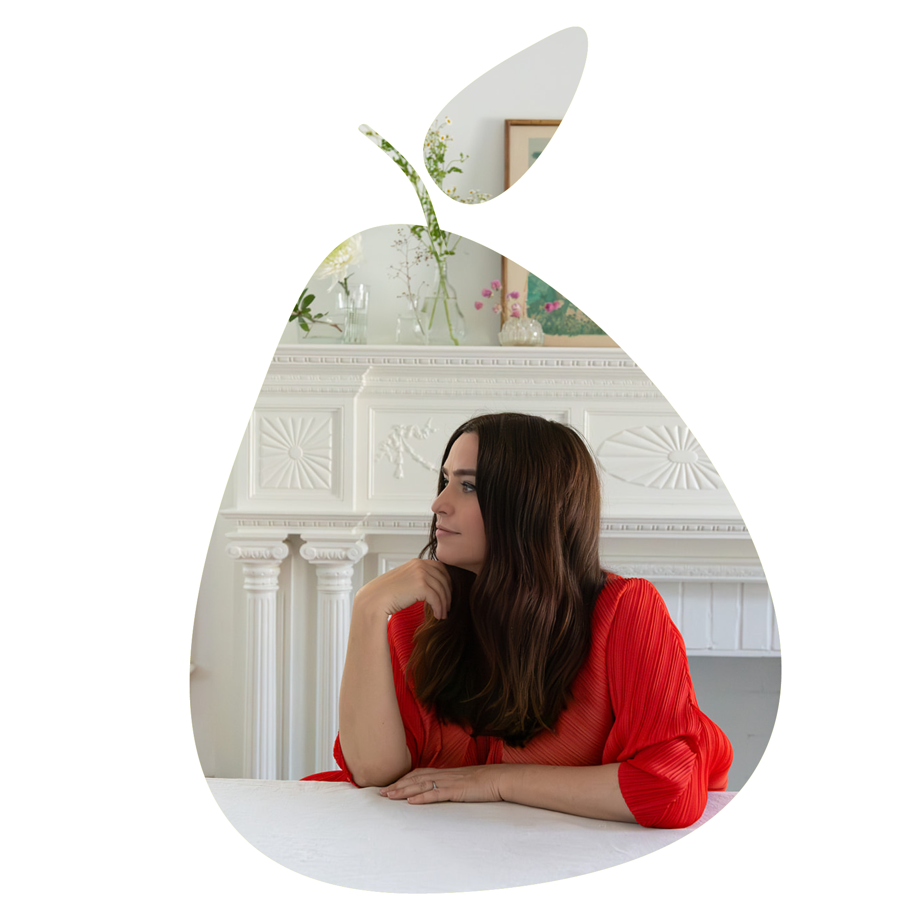 A woman with long brown hair in a red blouse sitting at a white table in a room with white ornate fireplace and decorated with vases of flowers and framed pictures.