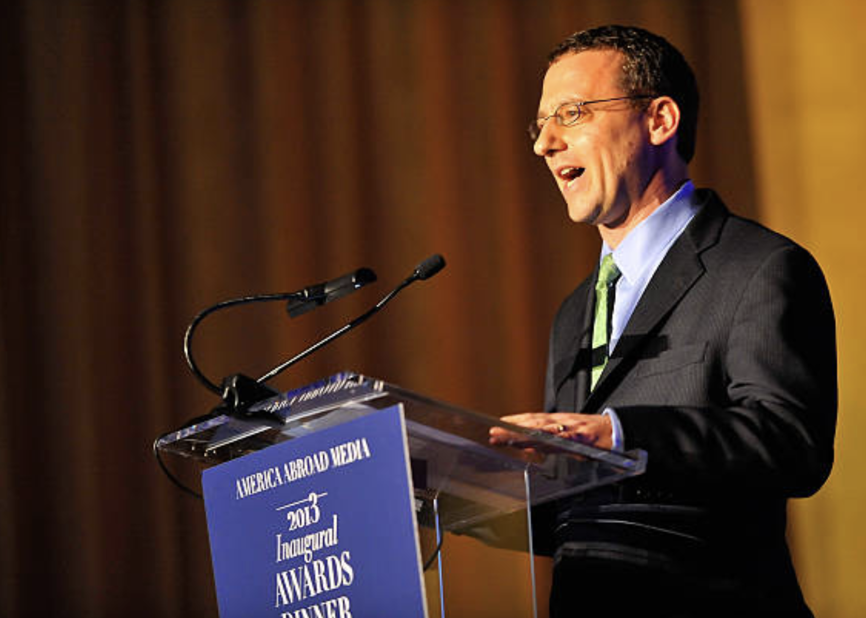 Man in a suit speaking at a podium with a sign that reads 'America Abroad Media 2013 Inaugural Awards Dinner'.