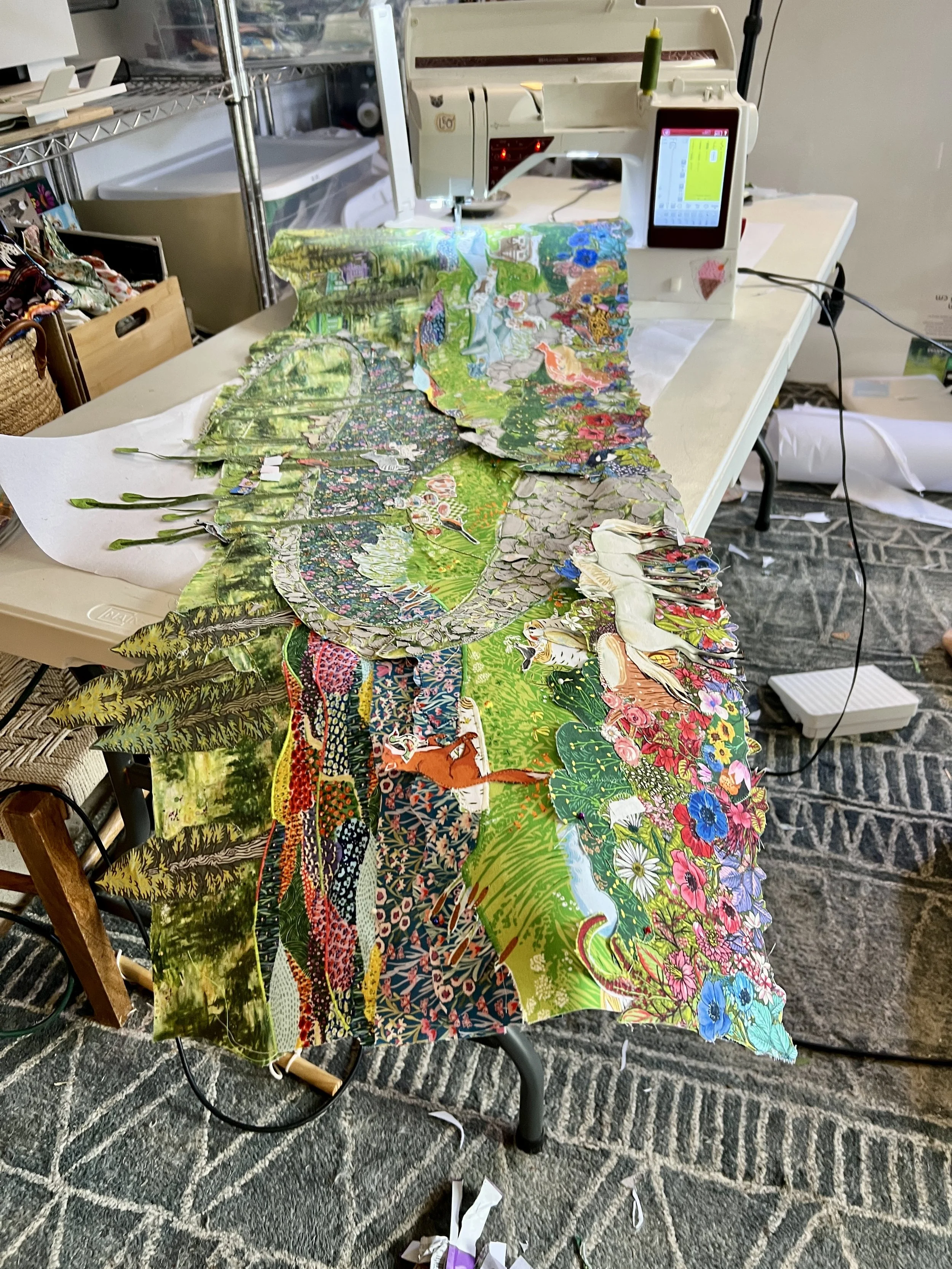 Colorful fabric with various nature and animal patterns laid out on a table in a sewing studio, with a sewing machine in the background.