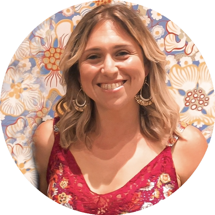 A woman with shoulder-length wavy blonde hair smiling in front of a floral patterned background, wearing a red floral dress and large earrings.