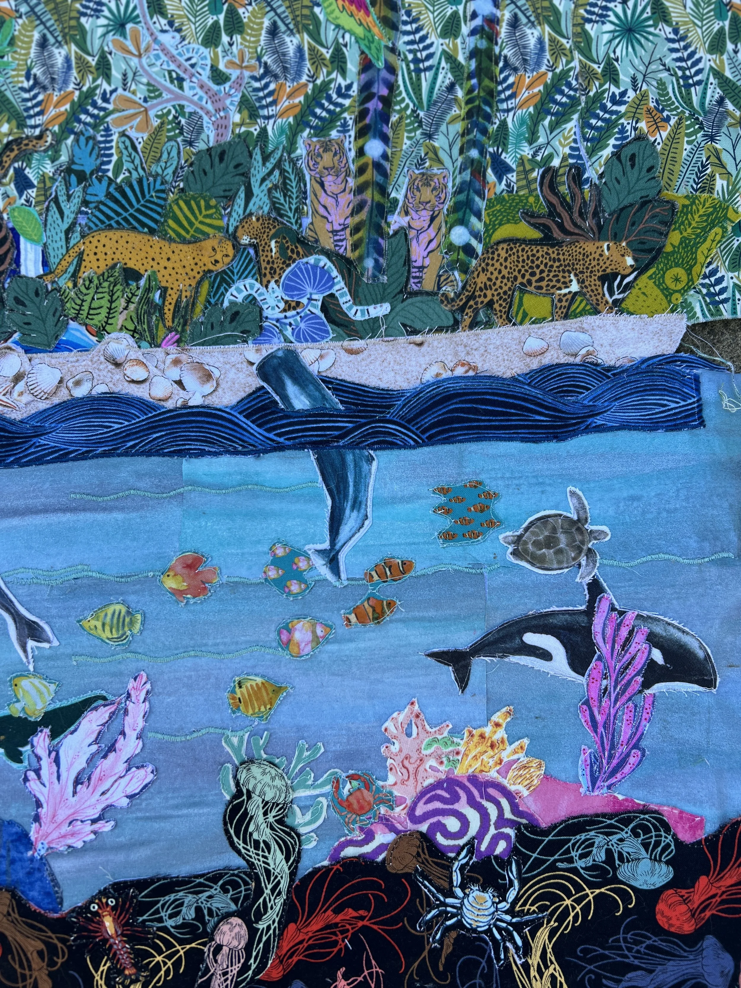 Colorful fabric art depicting a jungle scene with animals, water, and plants. The upper part shows leopards, jaguars, and other jungle animals among green foliage. The lower part illustrates underwater marine life, including a dolphin, sea turtle, fish, coral, and seashells.