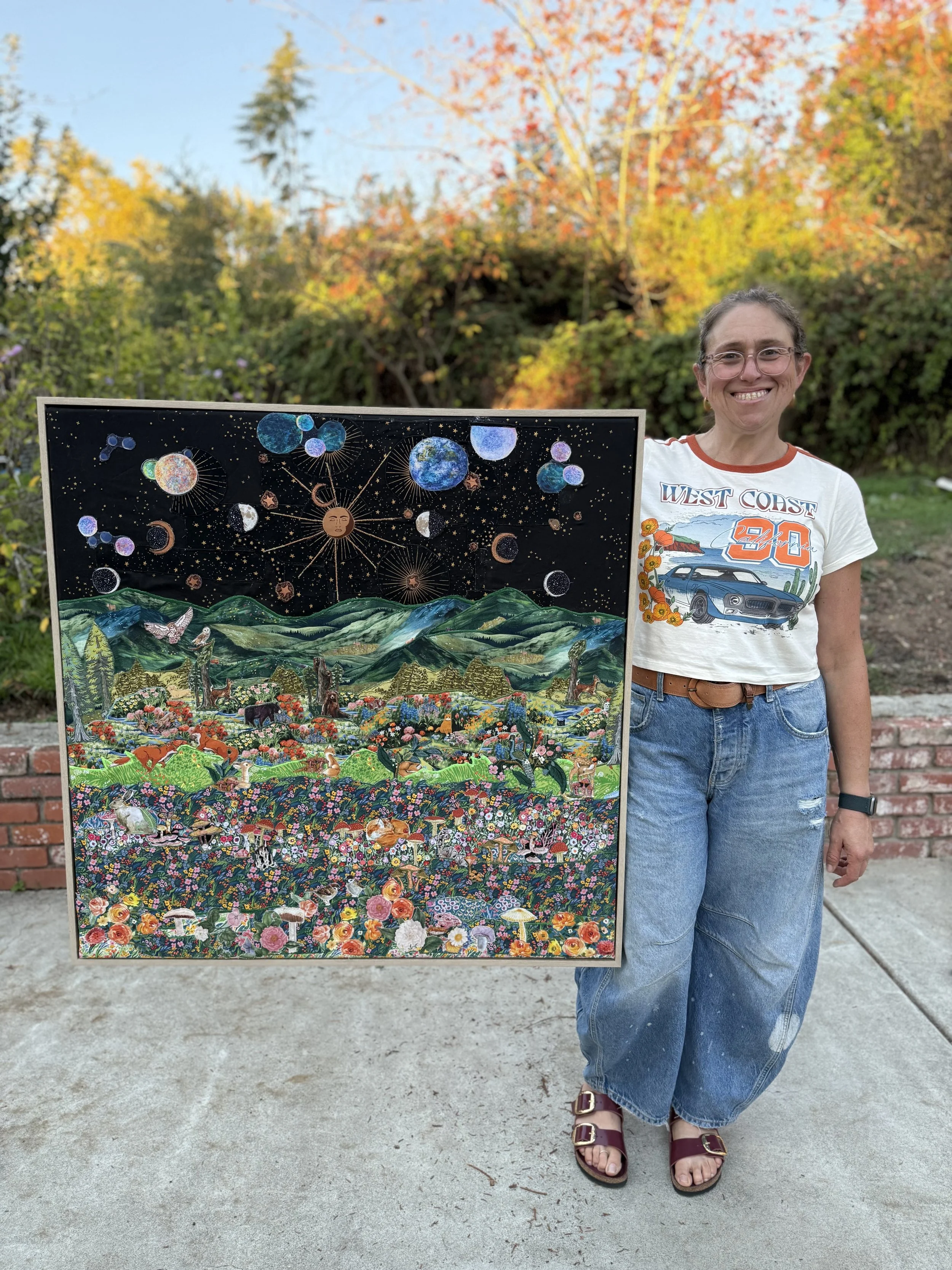 A woman stands outdoors in front of colorful fall trees, smiling while holding a large, vibrant, artistic collage featuring celestial objects above a lush green mountainous landscape filled with various animals and flowers.