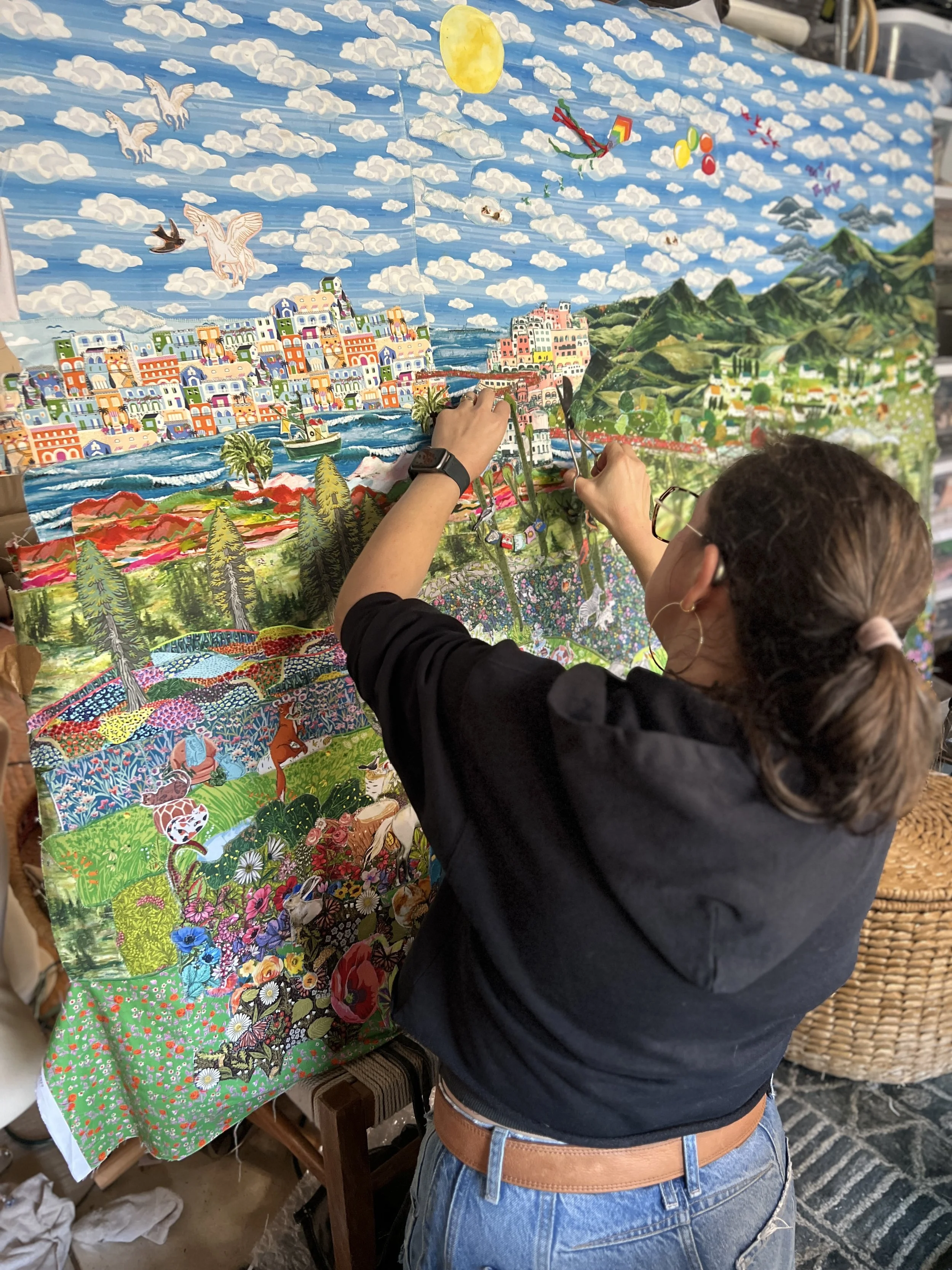 A woman with glasses and a black hoodie is working on a colorful, layered quilt or tapestry that depicts a scenic landscape with mountains, trees, animals, and a sky filled with clouds and a yellow sun.