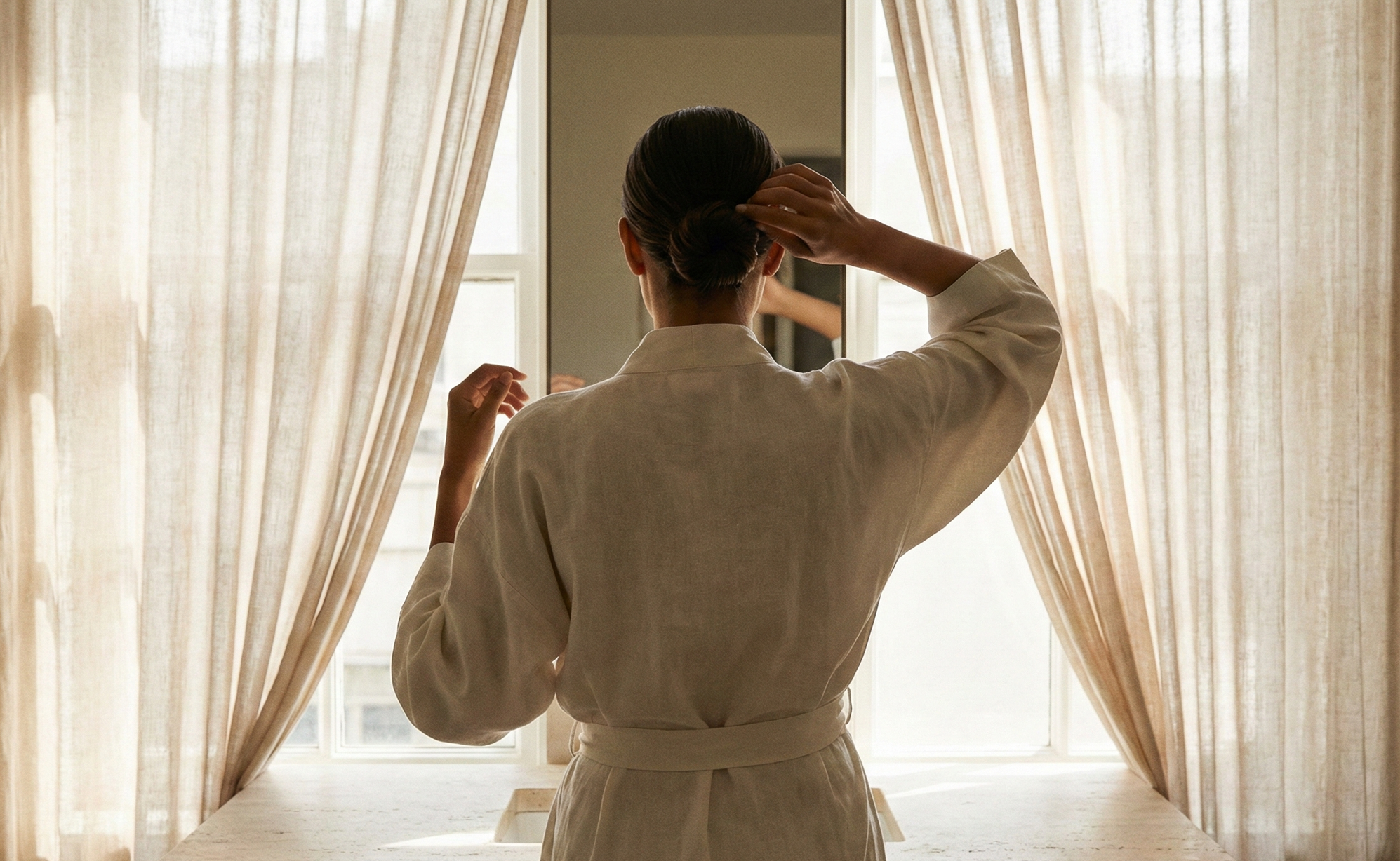 Back view of a woman in a white coat standing in front of a mirror near a window with beige curtains, adjusting her hair.