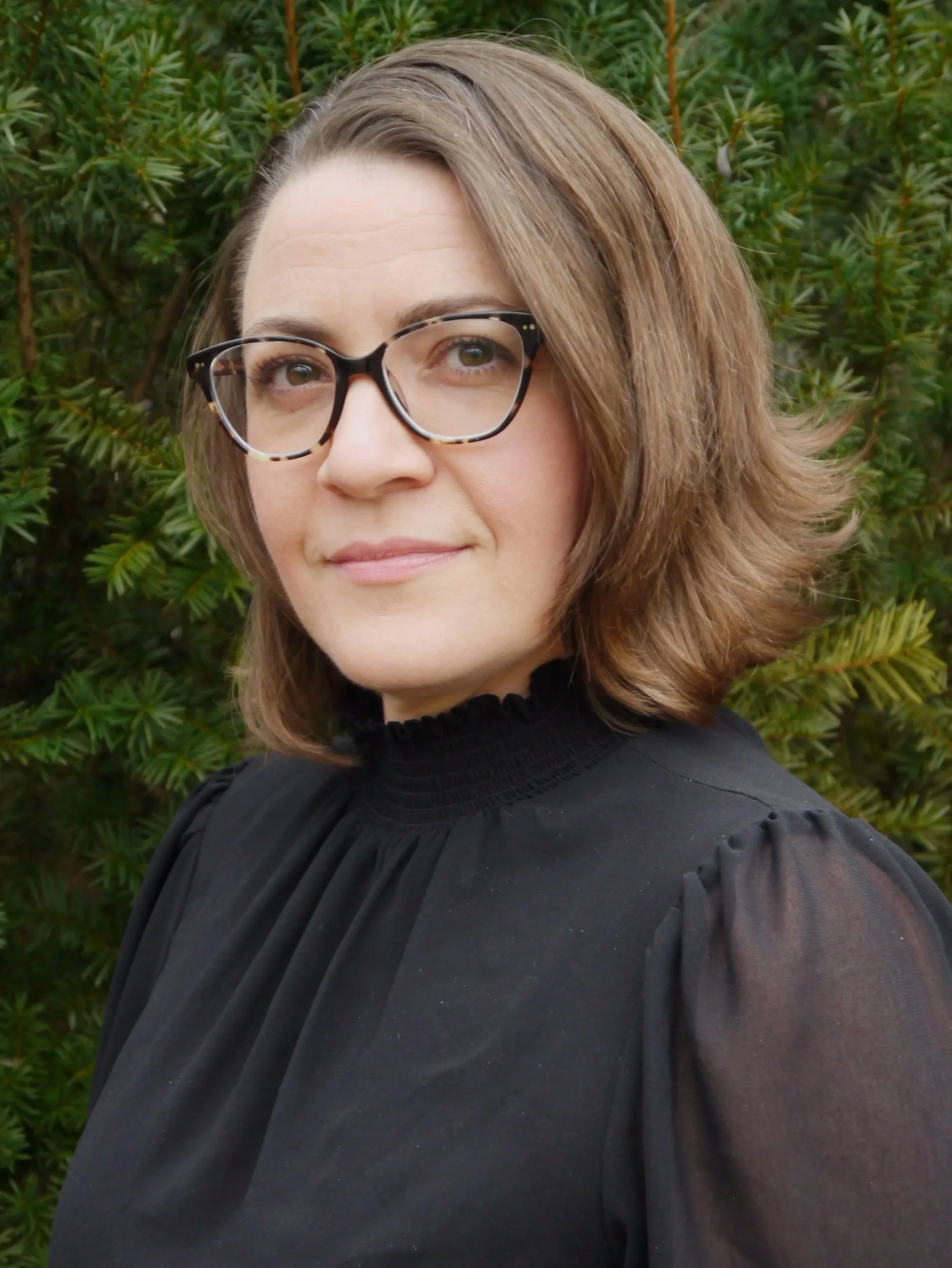 A woman with shoulder-length brown hair, wearing glasses, dressed in a black blouse, standing outdoors in front of green foliage.