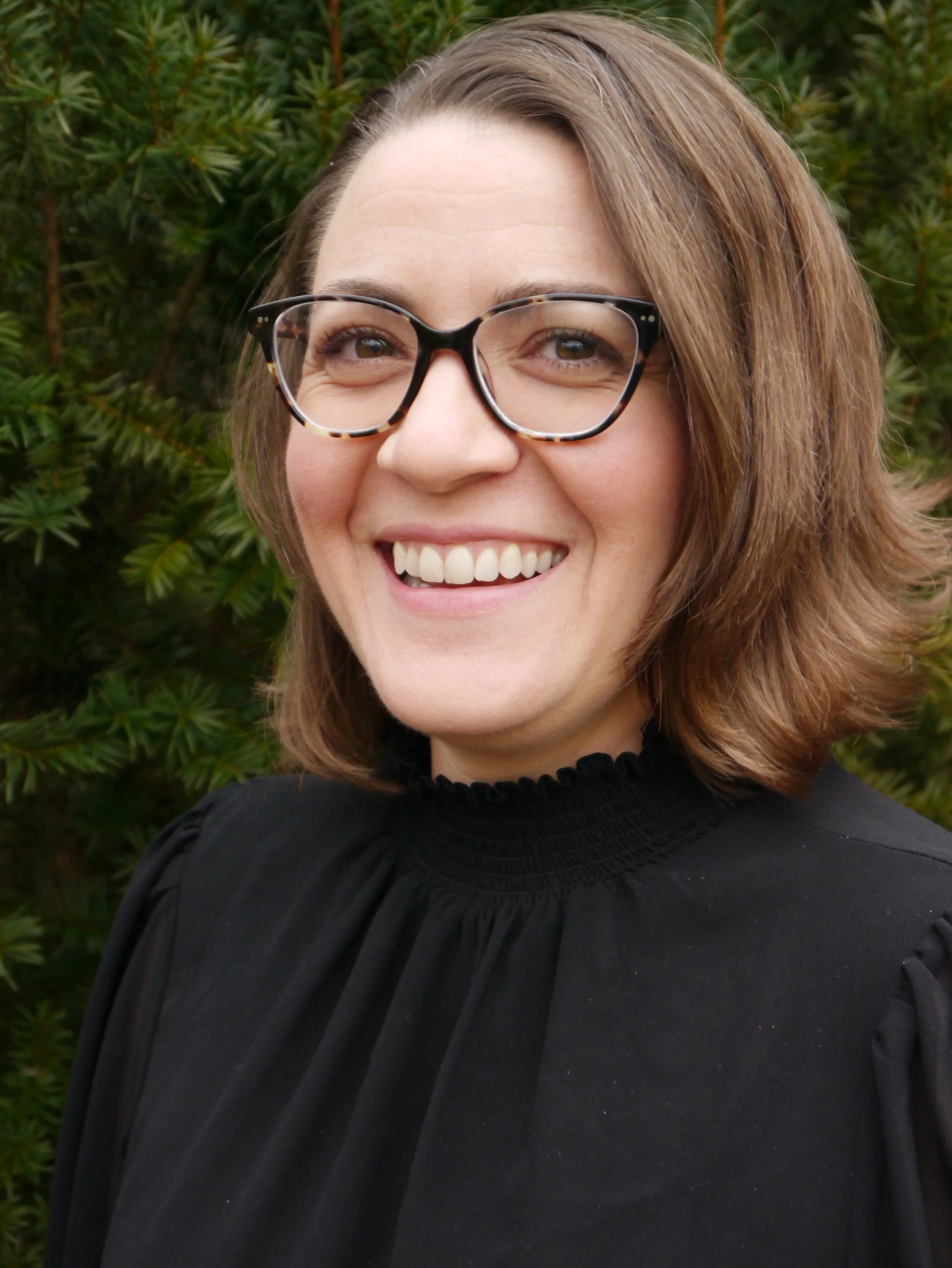 A smiling woman with shoulder-length brown hair, wearing glasses and a black blouse, standing outdoors with green foliage in the background.