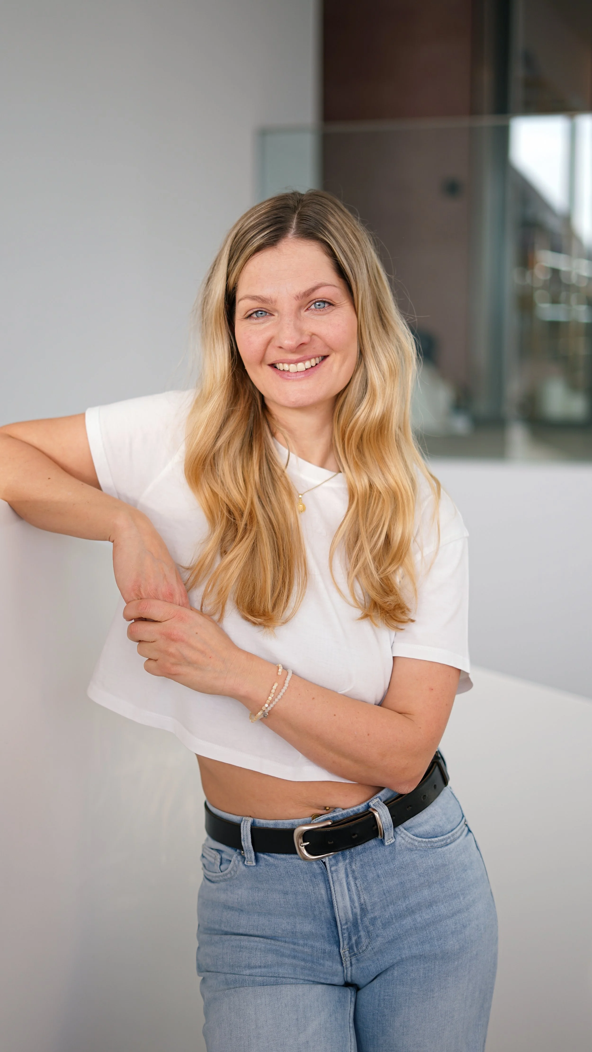 Junge Frau mit blonden, gewellten Haaren, lächelnd, trägt T-Shirt und Jeans, steht in einem modernen Raum.