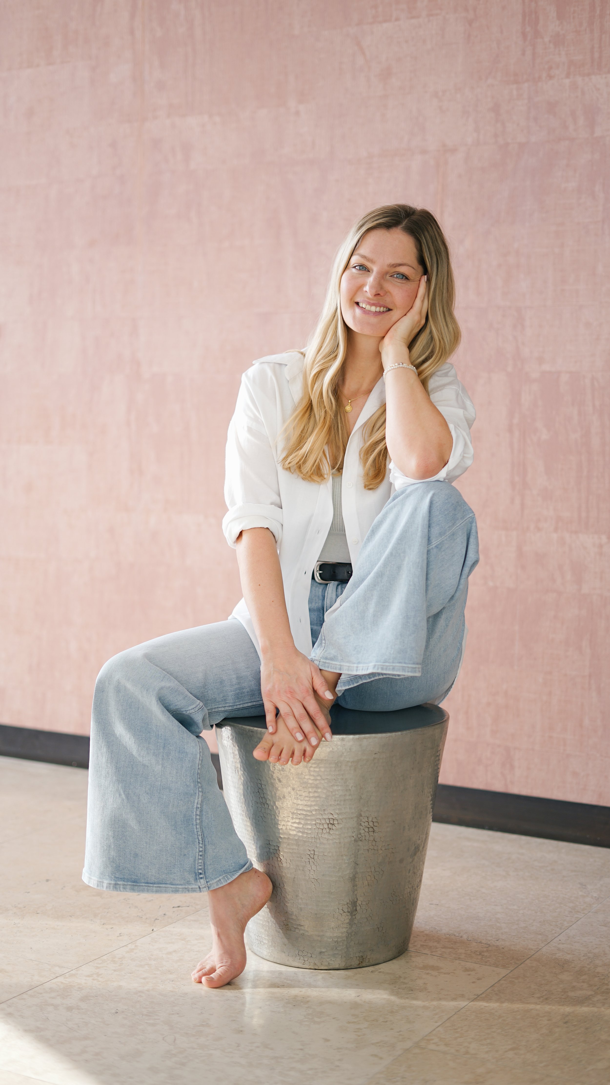 Eine lächelnde Frau sitzt barfuß auf einem Metallhocker vor einer rosa Wand, trägt weiße Bluse und Jeans.