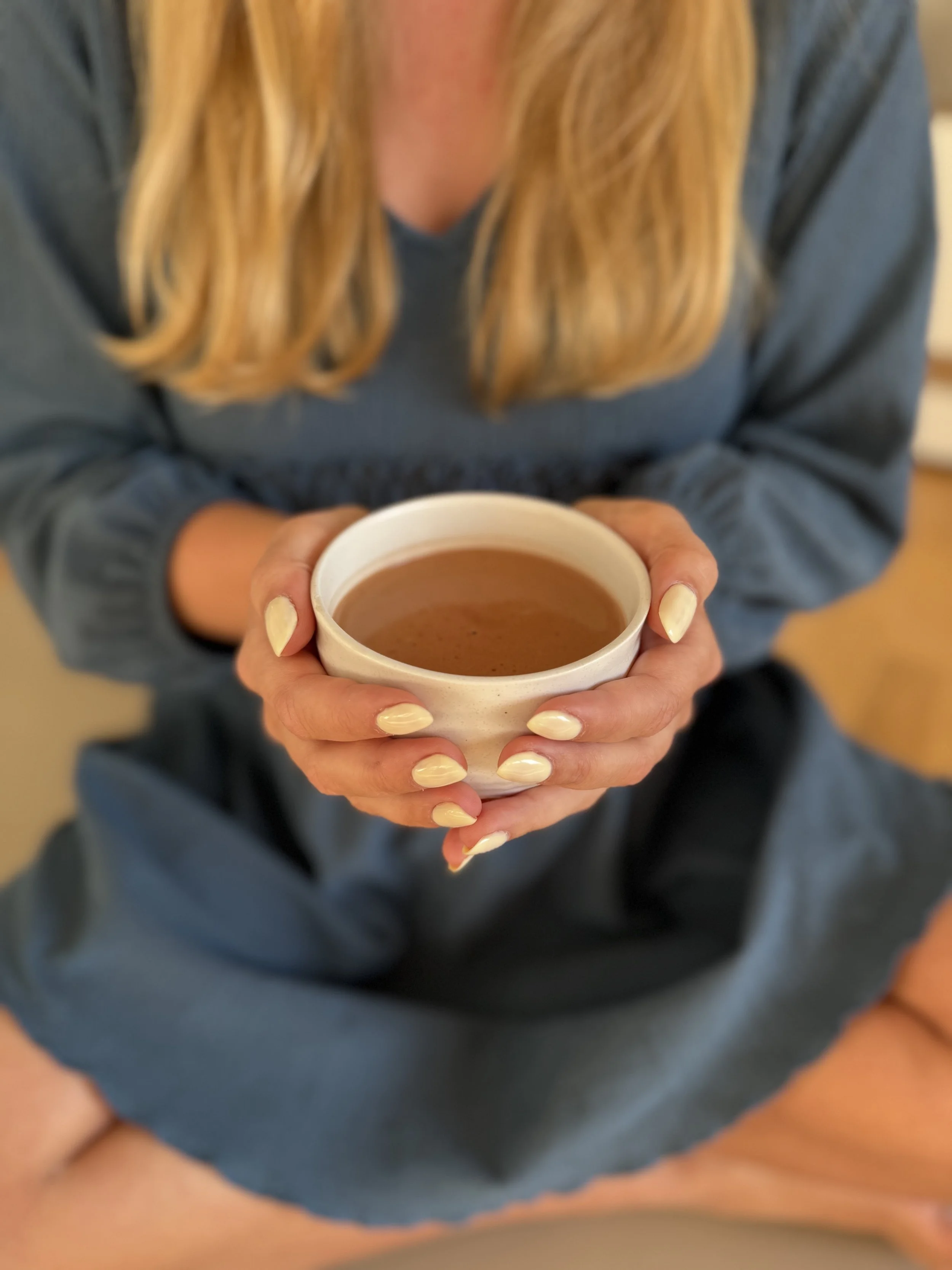 Frau hält eine Tasse Kaffee mit beiden Händen, Nägel lackiert, trägt einen blauen Pullover, sitzt auf dem Boden.