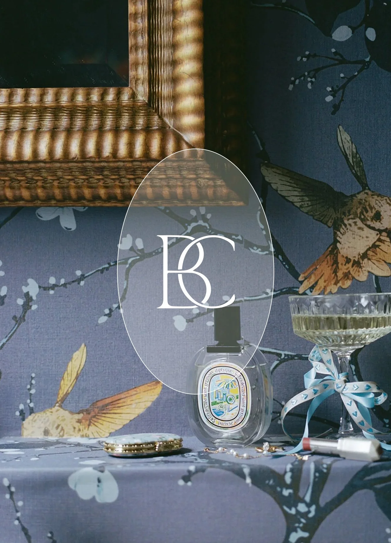 Decorative table setting with a textured gold mirror frame, a blue floral-patterned tablecloth with gold and yellow birds, a clear glass bowl with a ribbon, jewelry, and a bottle with a light label, all arranged on the table.
