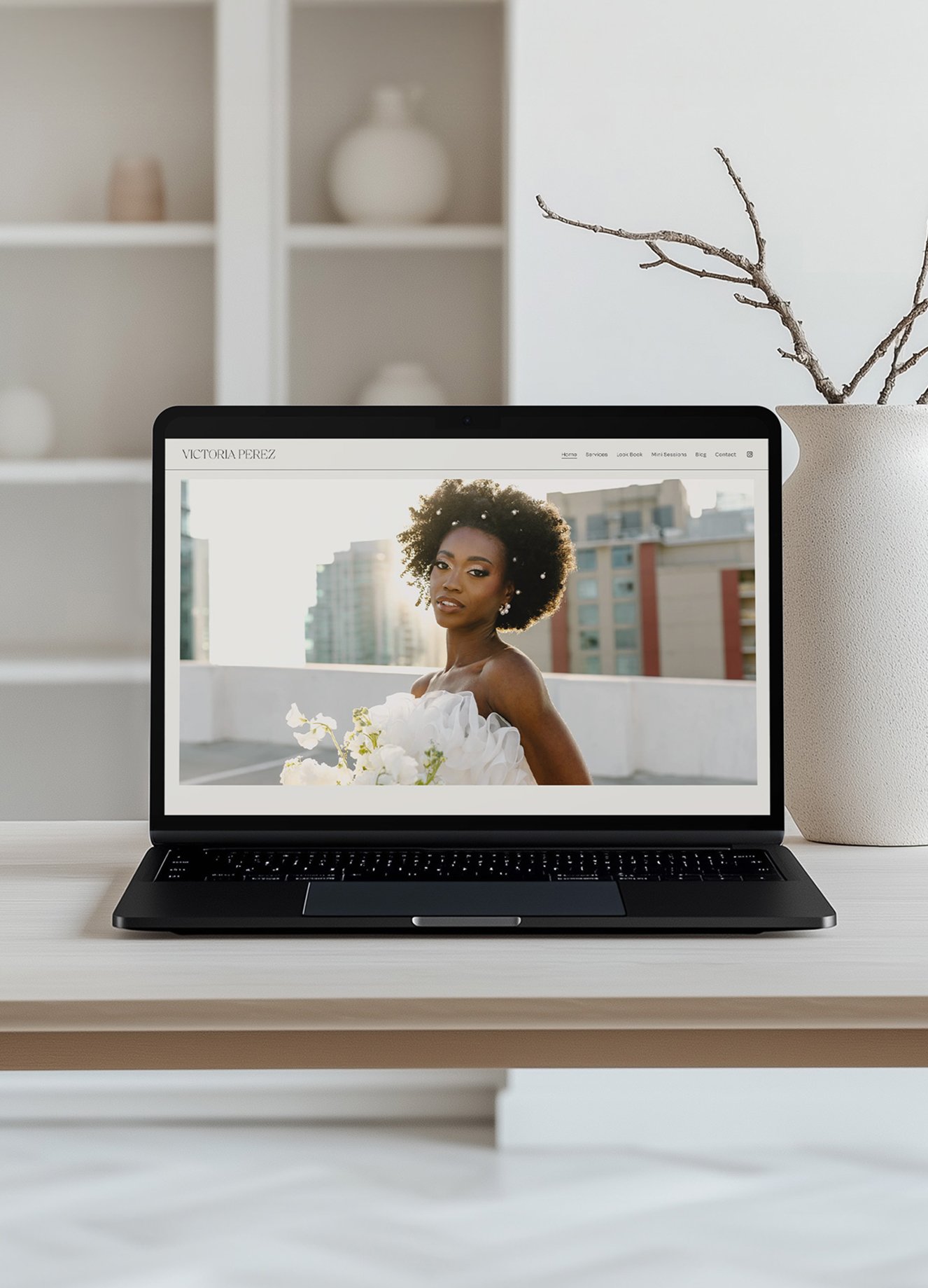 A laptop on a white desk displaying a portrait of a woman with natural hair, holding white flowers. A beige vase with tree branches is to the right, and a white bookshelf with decorative objects is in the background.