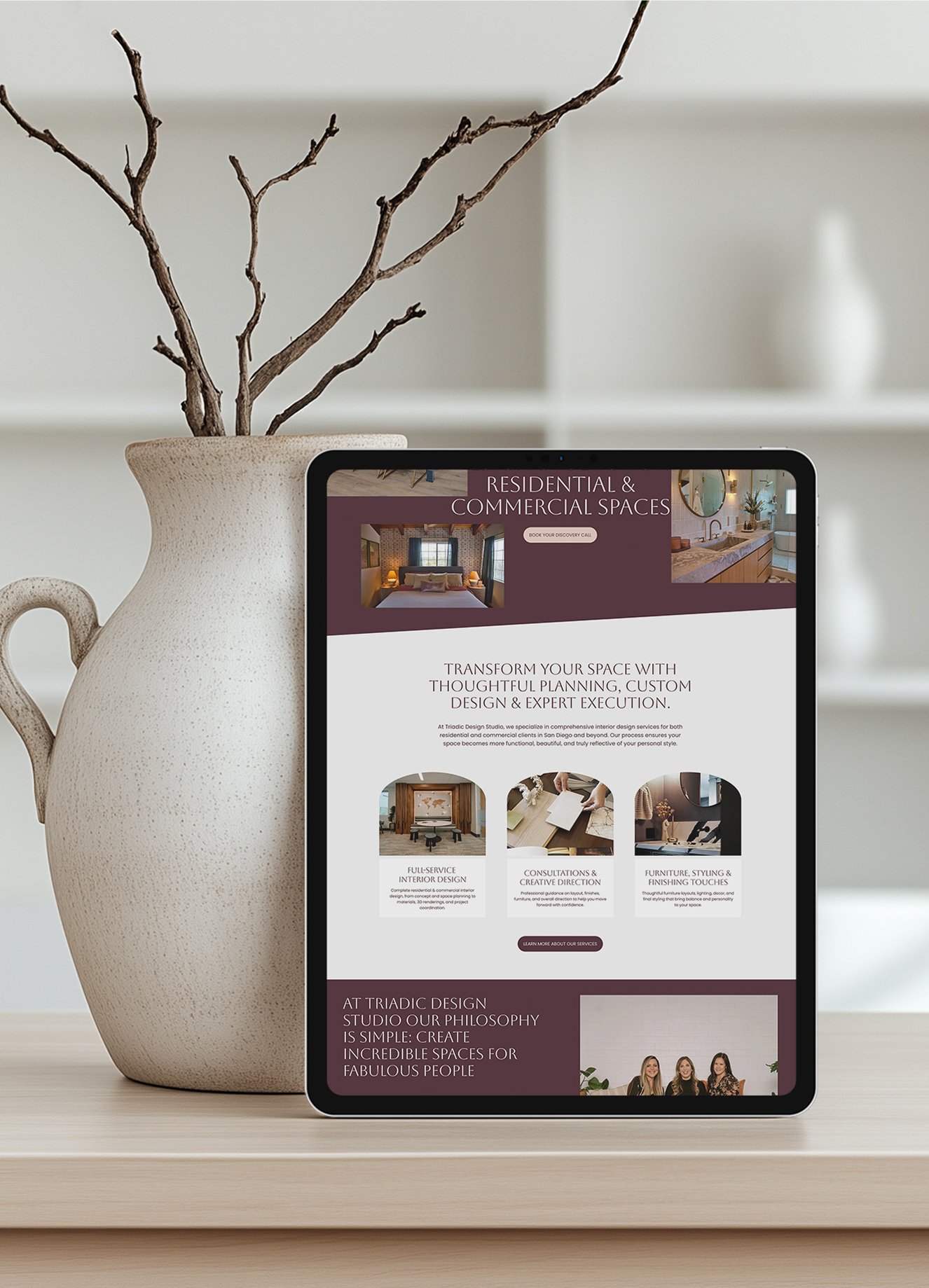 A digital tablet displaying a website for Triadic Design Studio with interior design services. The tablet is placed on a light-colored surface, next to a beige ceramic vase with twisted tree branches inside. The background includes a white shelf and a blurred white vase.