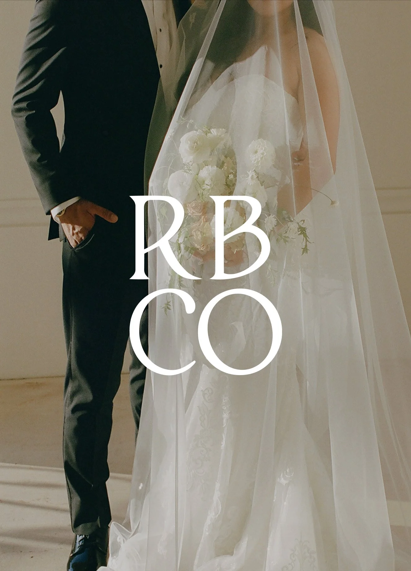 A bride and groom standing together at their wedding, with the bride in a white wedding gown holding a bouquet and the groom in a black suit with his hand in his pocket. There is a large text over the image that says "RBCO".
