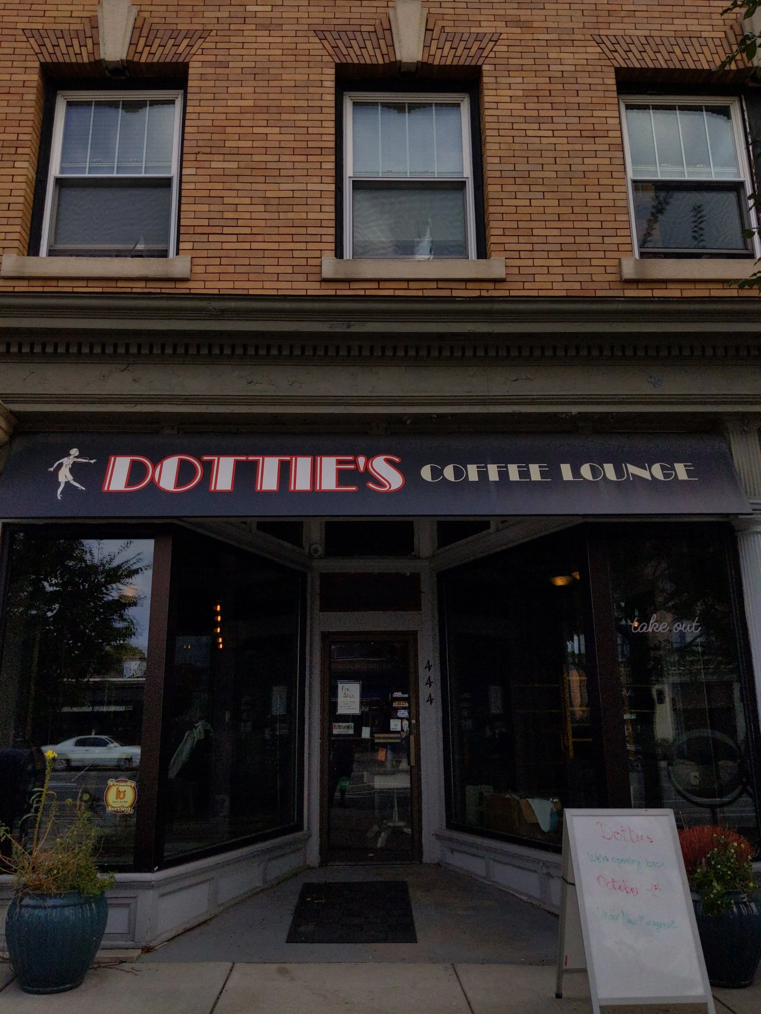 Exterior of Dottie's Coffee Lounge with a black sign and red-white lettering, brick building, and glass windows, with plants on either side of the entrance and a whiteboard sign outside.