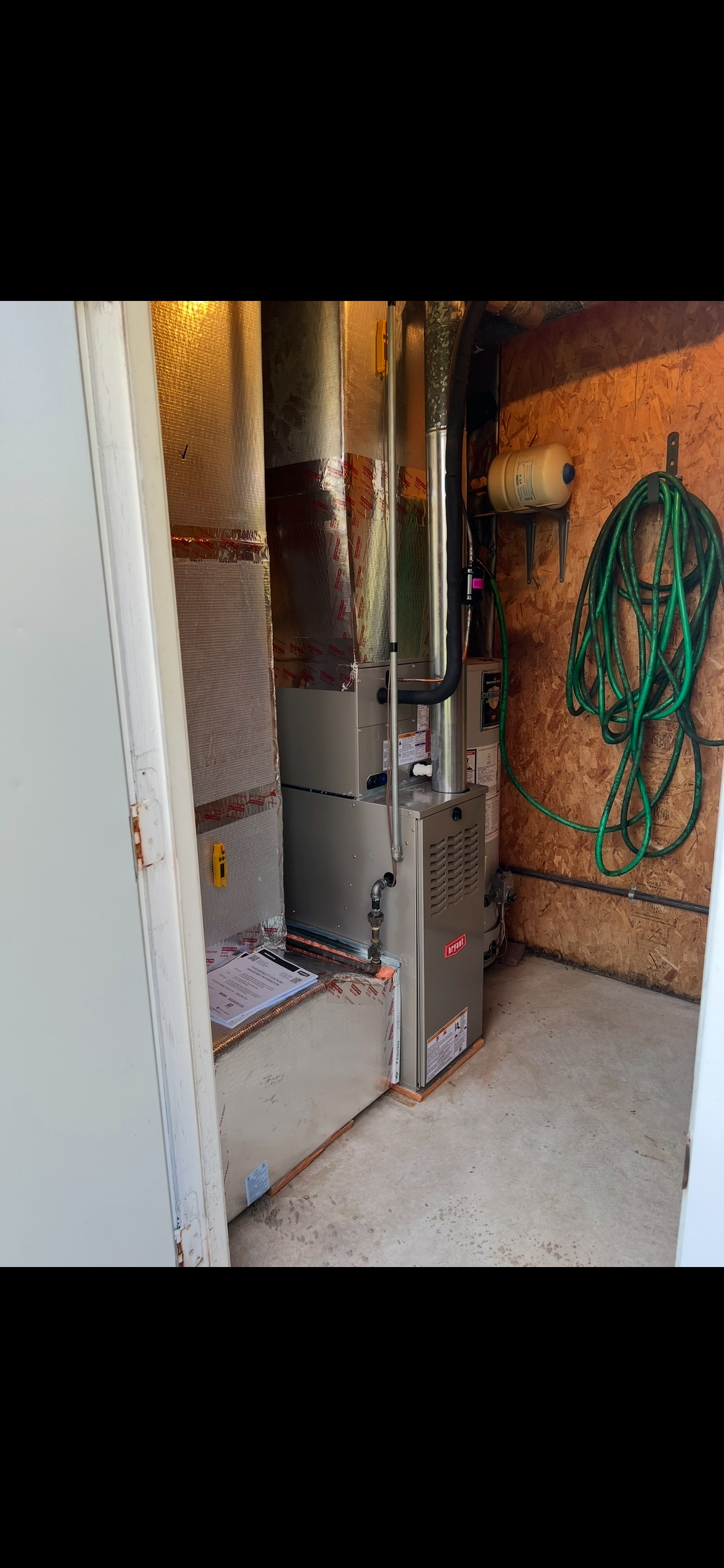 HVAC furnace unit installed in a utility room with insulated ductwork, a coiled garden hose, and a propane tank.