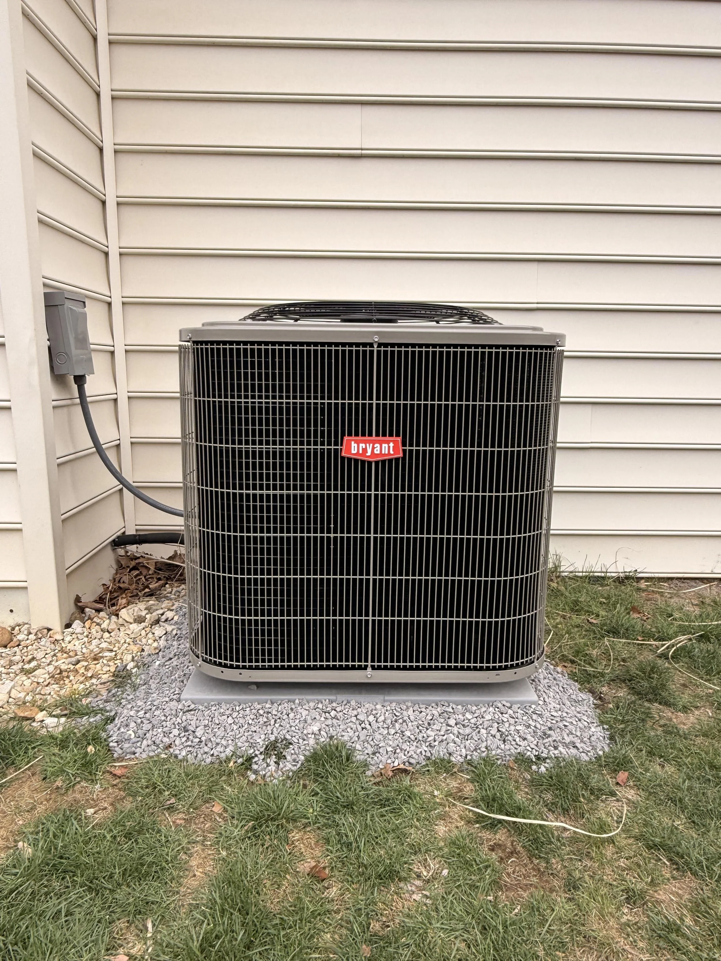 Outdoor air conditioning unit with a Bryant logo, placed on a small gravel pad, with a beige siding house wall in the background and some patchy grass and leaves on the ground.