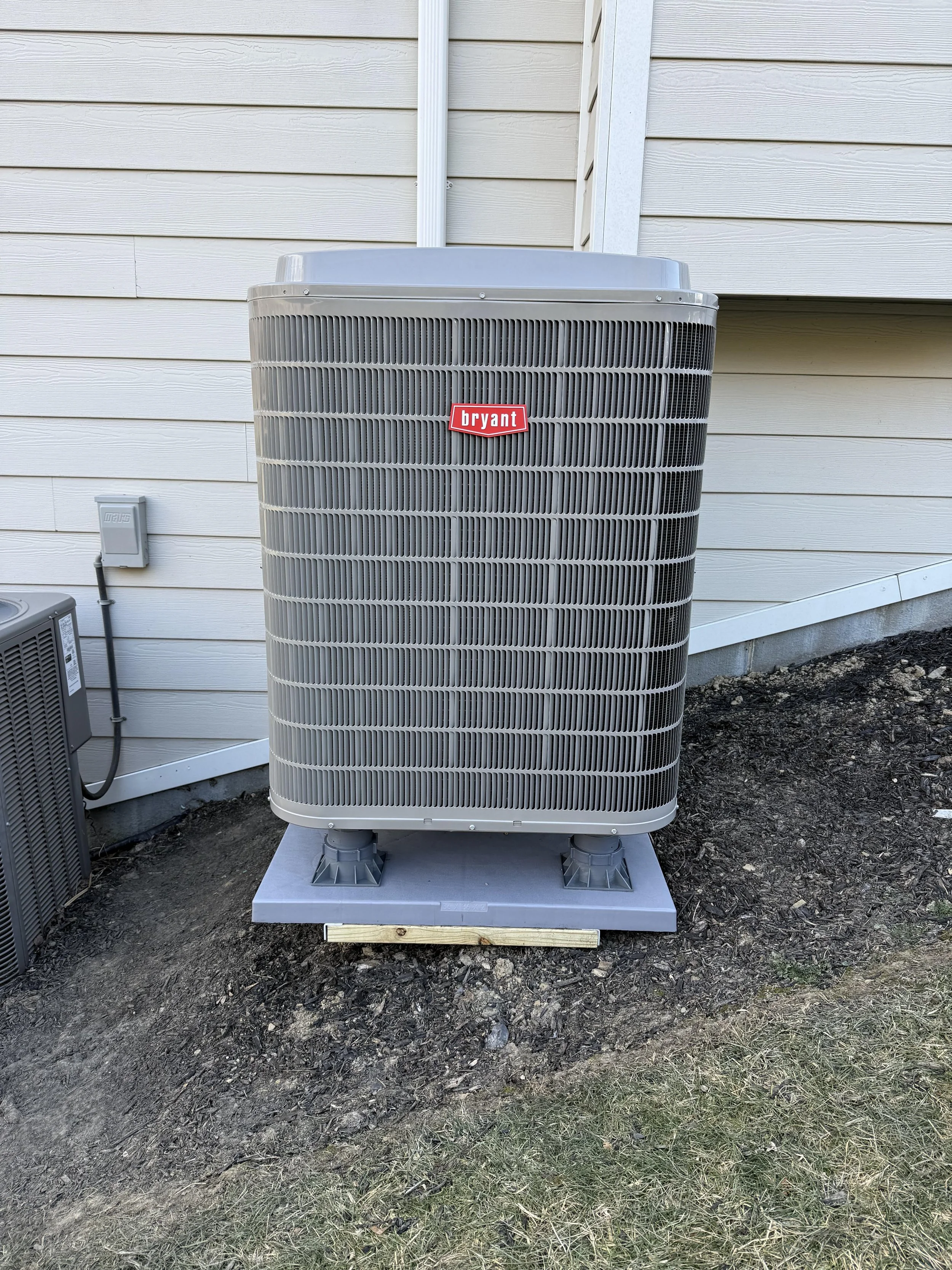 An outdoor Bryant air conditioning unit installed on a concrete pad against the side of a house with beige vinyl siding. There is a smaller gray unit nearby and an electrical outlet on the wall.