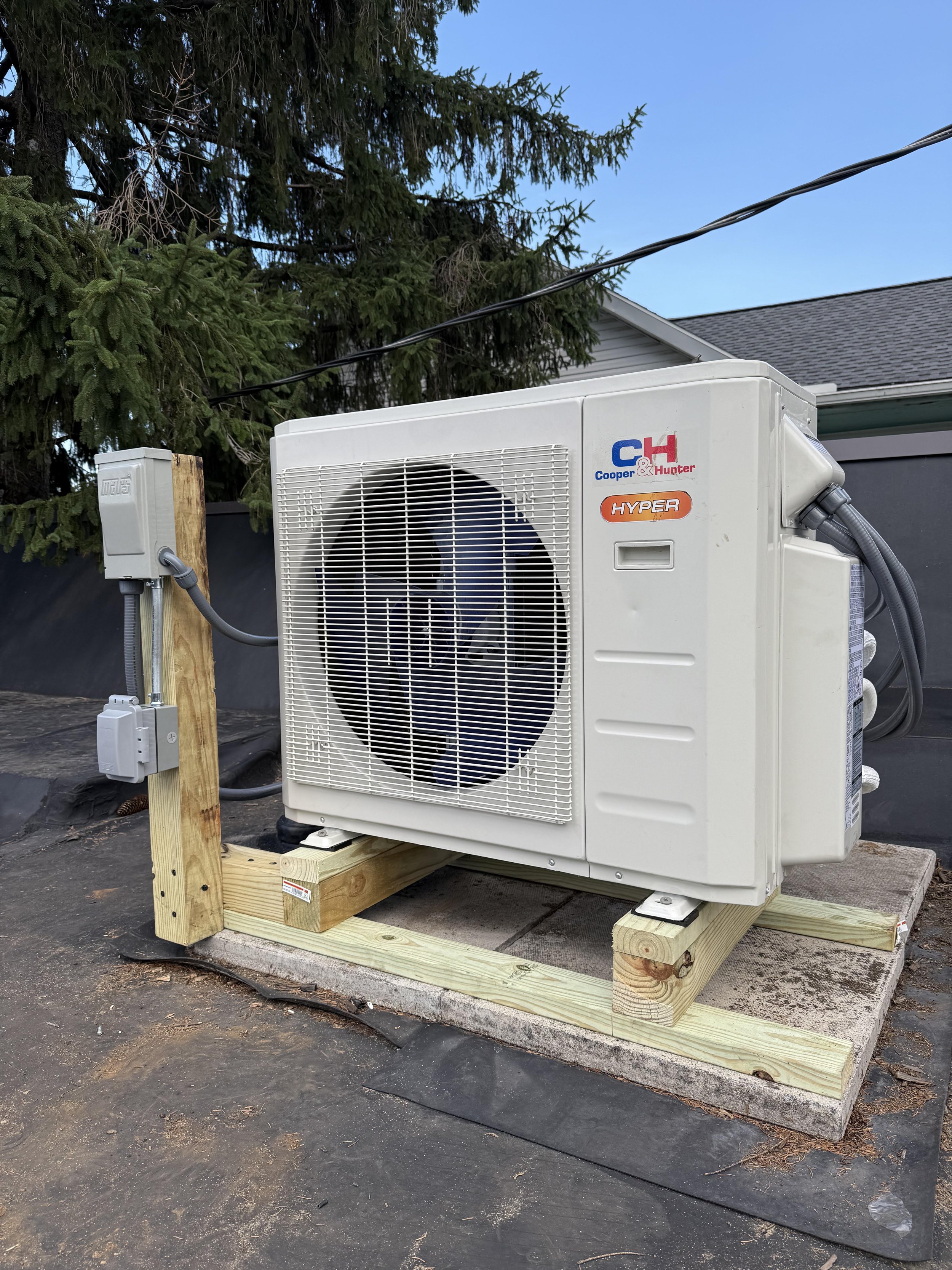 An outdoor air conditioning unit mounted on a wooden frame with a large pine tree in the background.