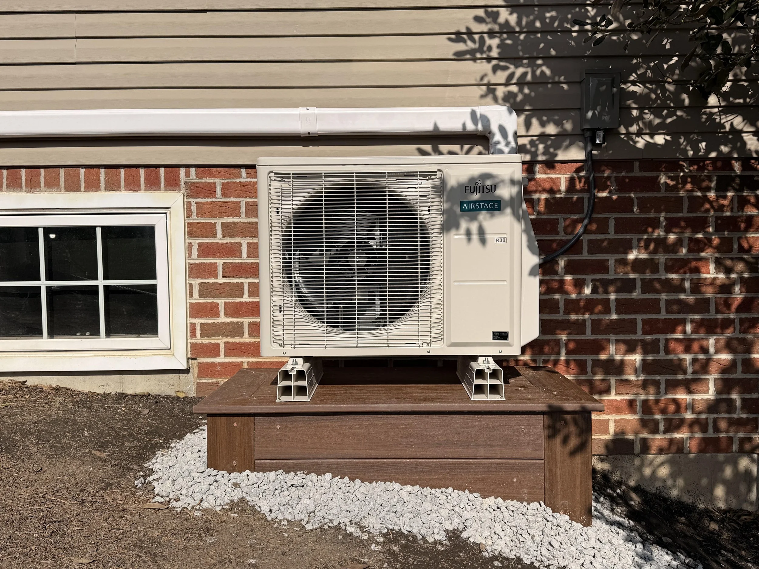 Exterior wall with beige siding, red brick foundation, white-framed window, and Fujitsu Airstage HVAC unit on a brown wooden platform, cast shadows from tree branches.