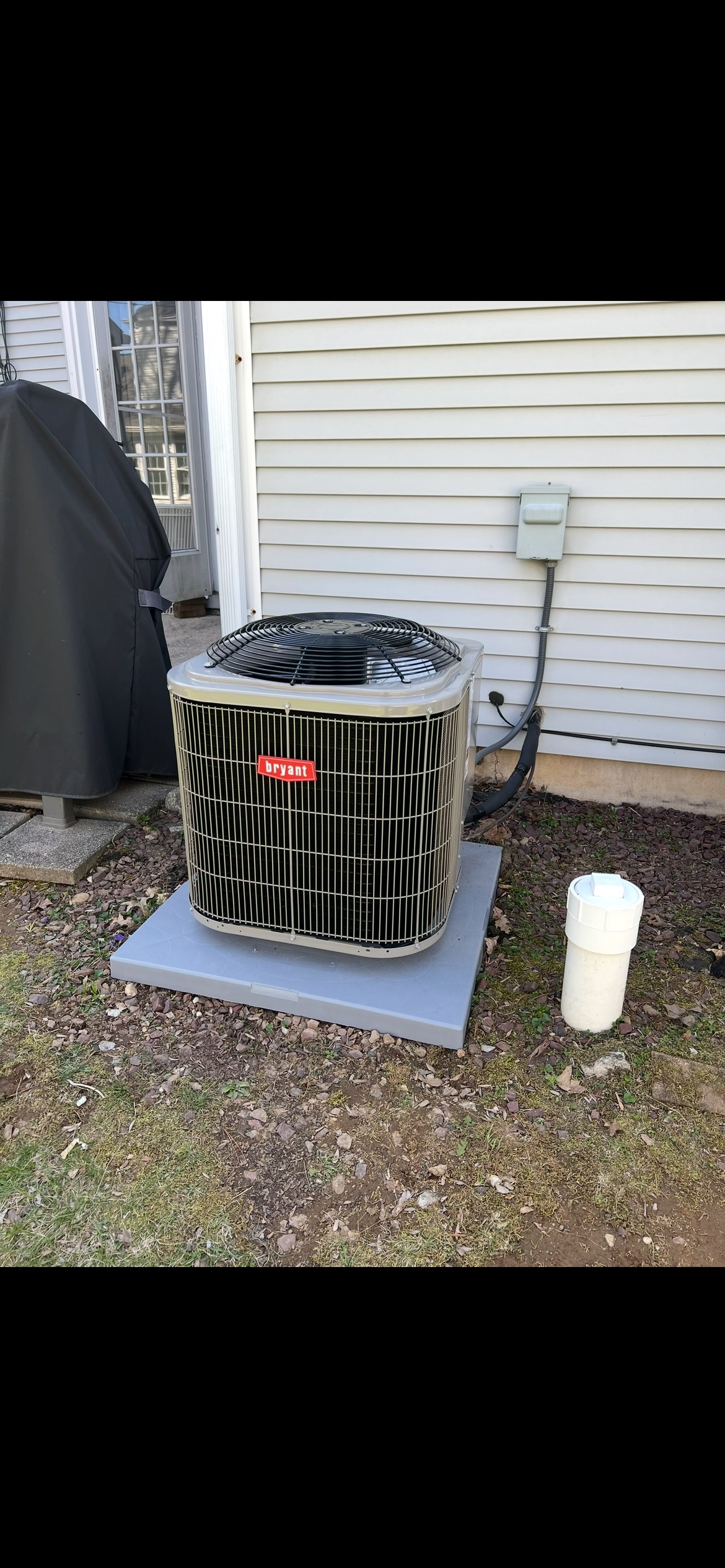 Bryant air conditioning unit outside a house on a concrete pad, with a covered barbecue grill to the left and a white utility box and drainage pipe on the wall to the right.