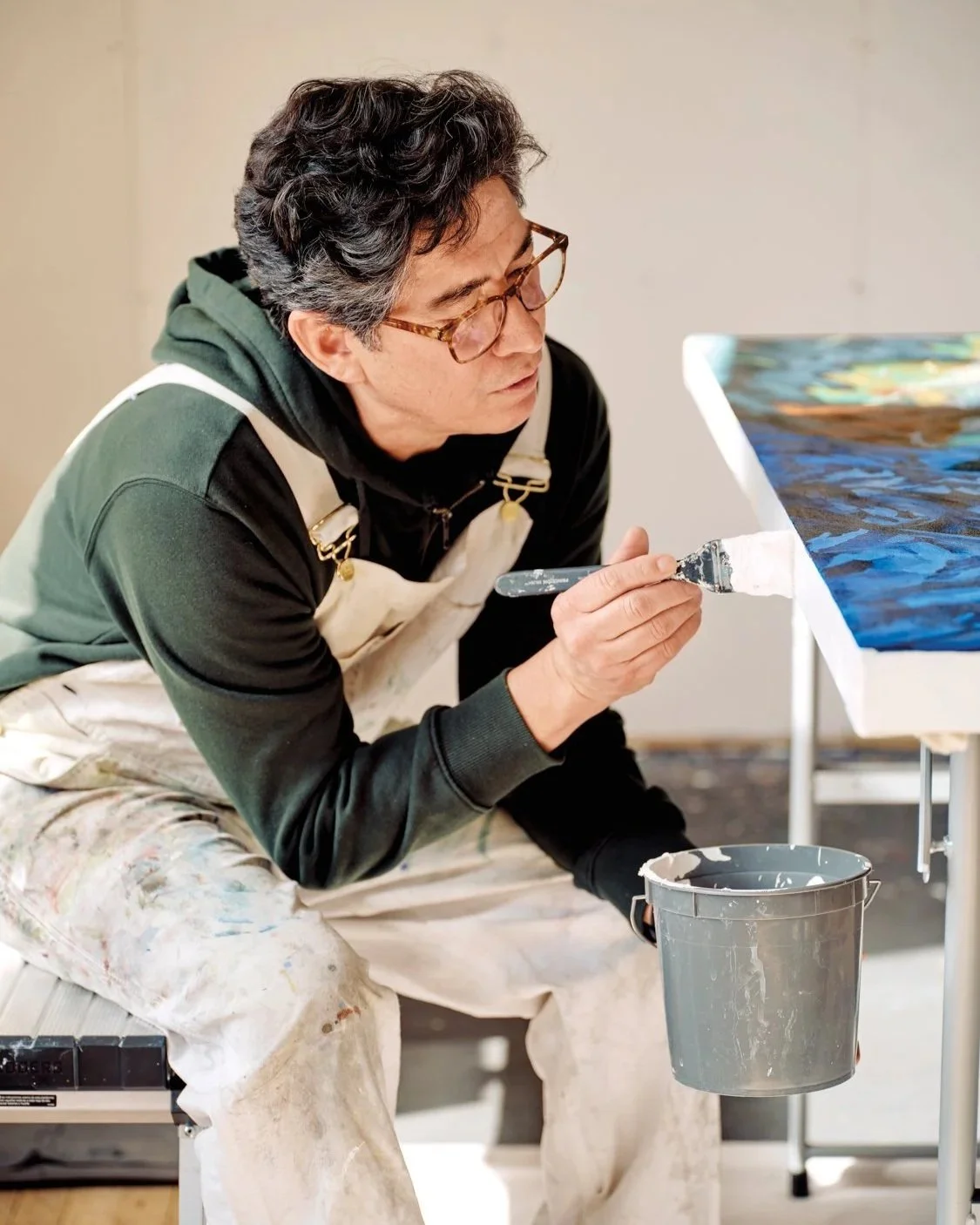 Steven J. Yazzie painting in his studio wearing glasses, a black hoodie, and white overalls with paint stains, sitting and painting a landscape on a canvas.