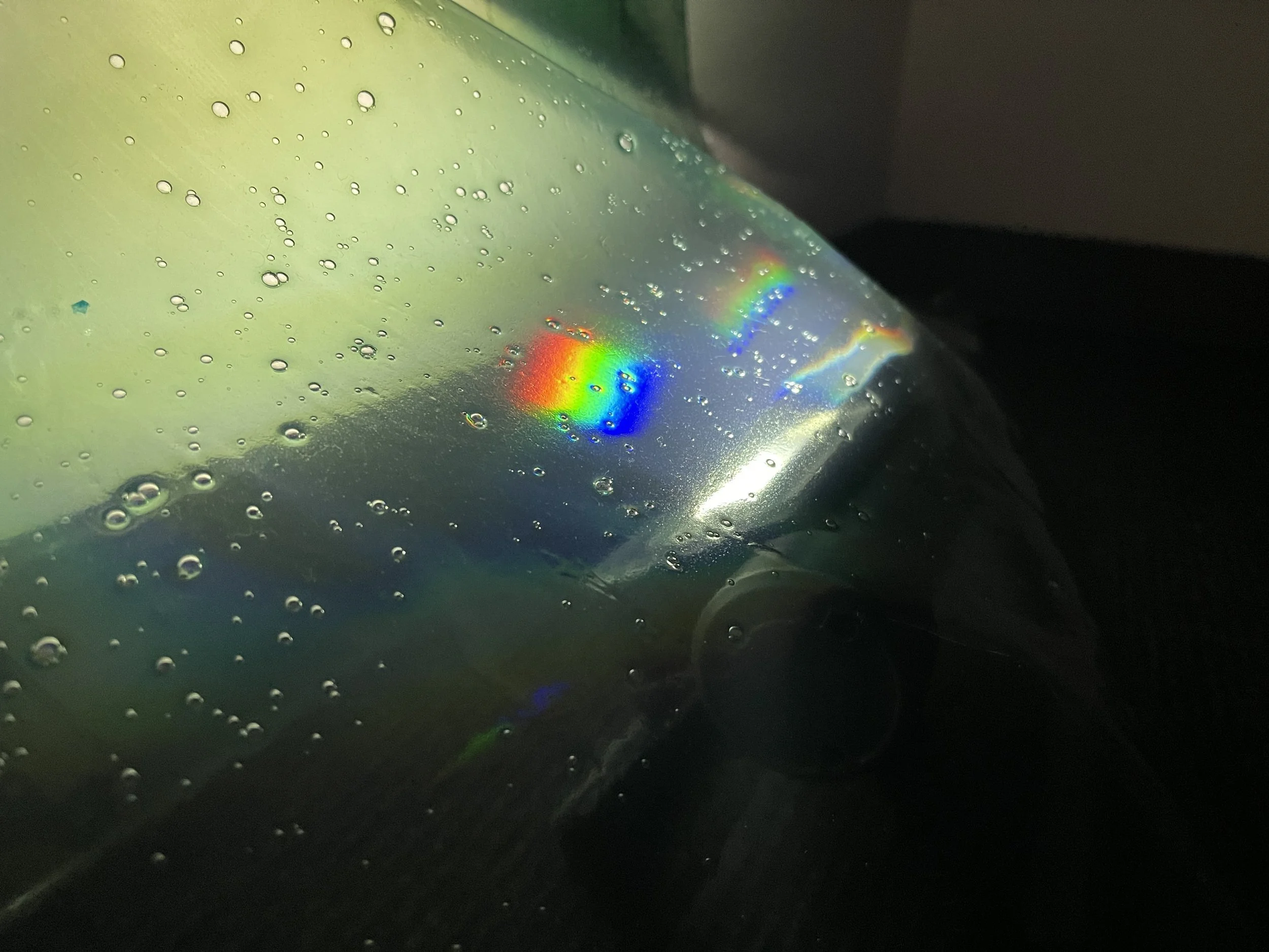 A photo by Janna Avner Close-up of a CD with water droplets on its surface, reflecting rainbow colors from light. Janna Avner (Koyukon Athabascan) and Sasha Fishman, "Bioplastic (Blooper Prism) detail", 2021. Chitosan, algae, hologram, LED light, 20"