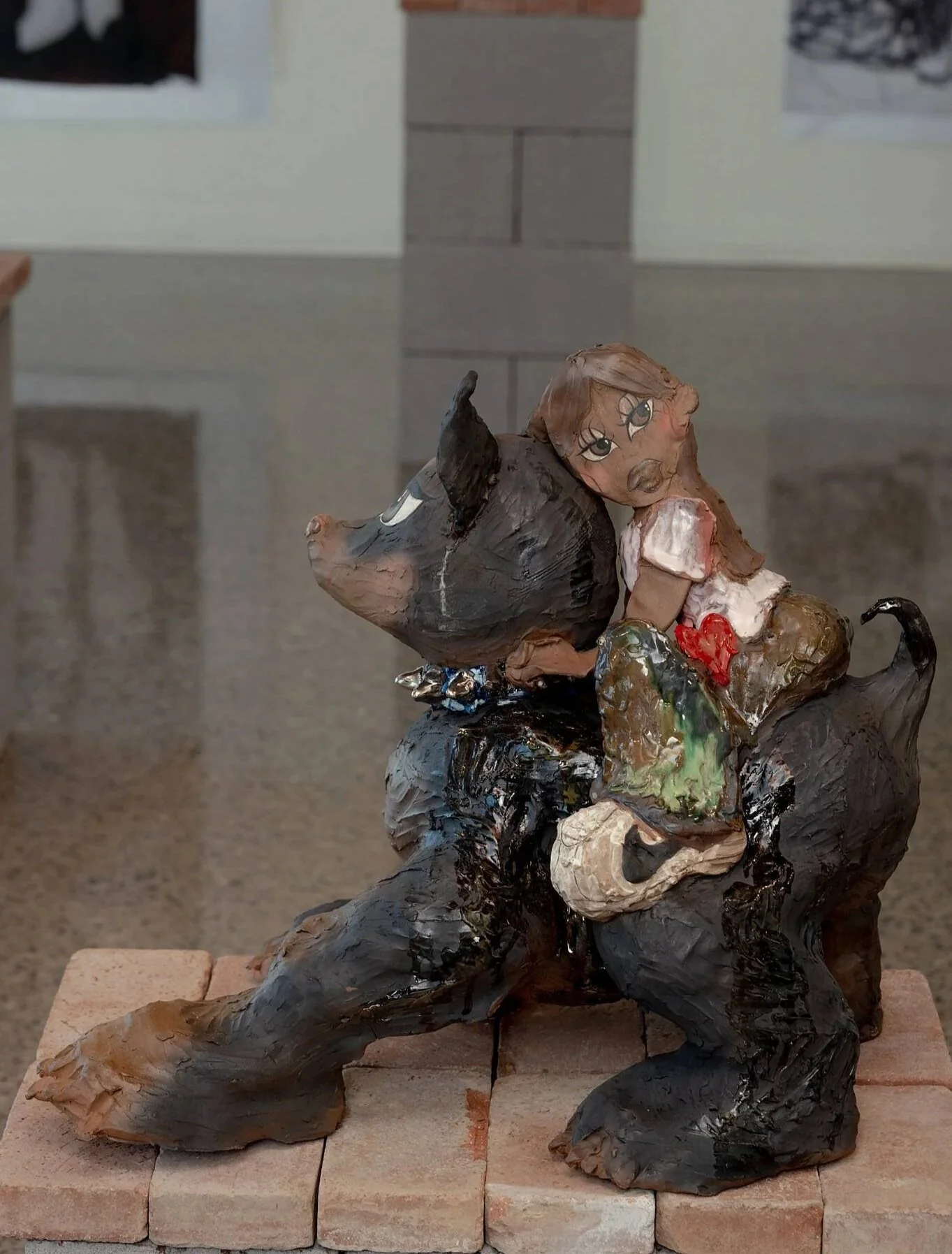 Stoneware sculpture by Yaritza Flores Bustos of a girl with long hair and large eyes sitting on a black dog, holding a red heart-shaped object. The sculpture is painted with earthy colors and is placed on a brick platform.