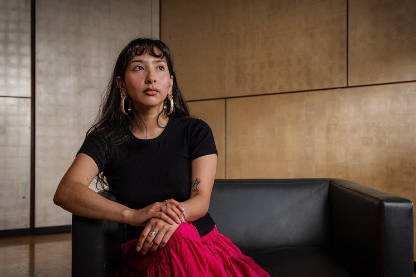 Yaritza Flores Bustos sitting on a black leather couch in a room with wooden walls at the Phoenix Art Museum for an interview about being a Sally and Richard Lehmann Emerging Artist Award recipient.
