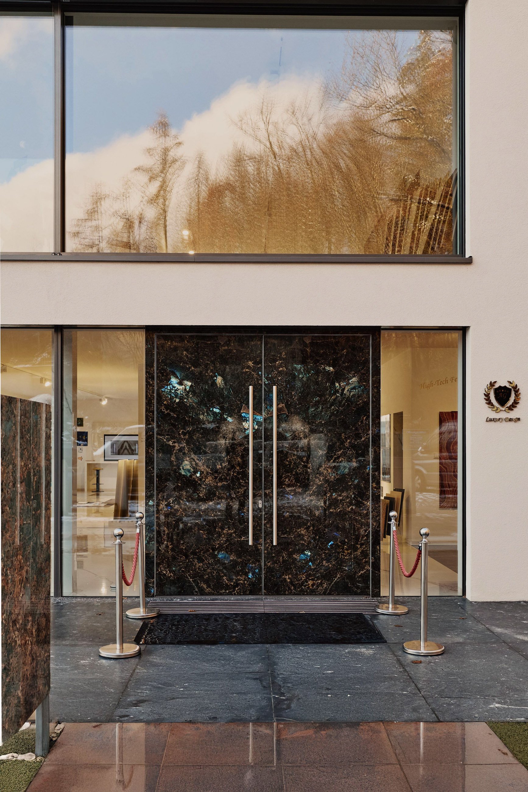 Der Eingangsbereich eines modernen Gebäudes mit schwarzen Marmortüren, seitlichen Glaspaneelen und einer weißen Fassade. Es gibt eine schwarze Matte vor den Türen und eine Absperrung aus Ständern mit rotem Samtseil. Im Inneren sind einige Bilder und Beleuchtung sichtbar.
