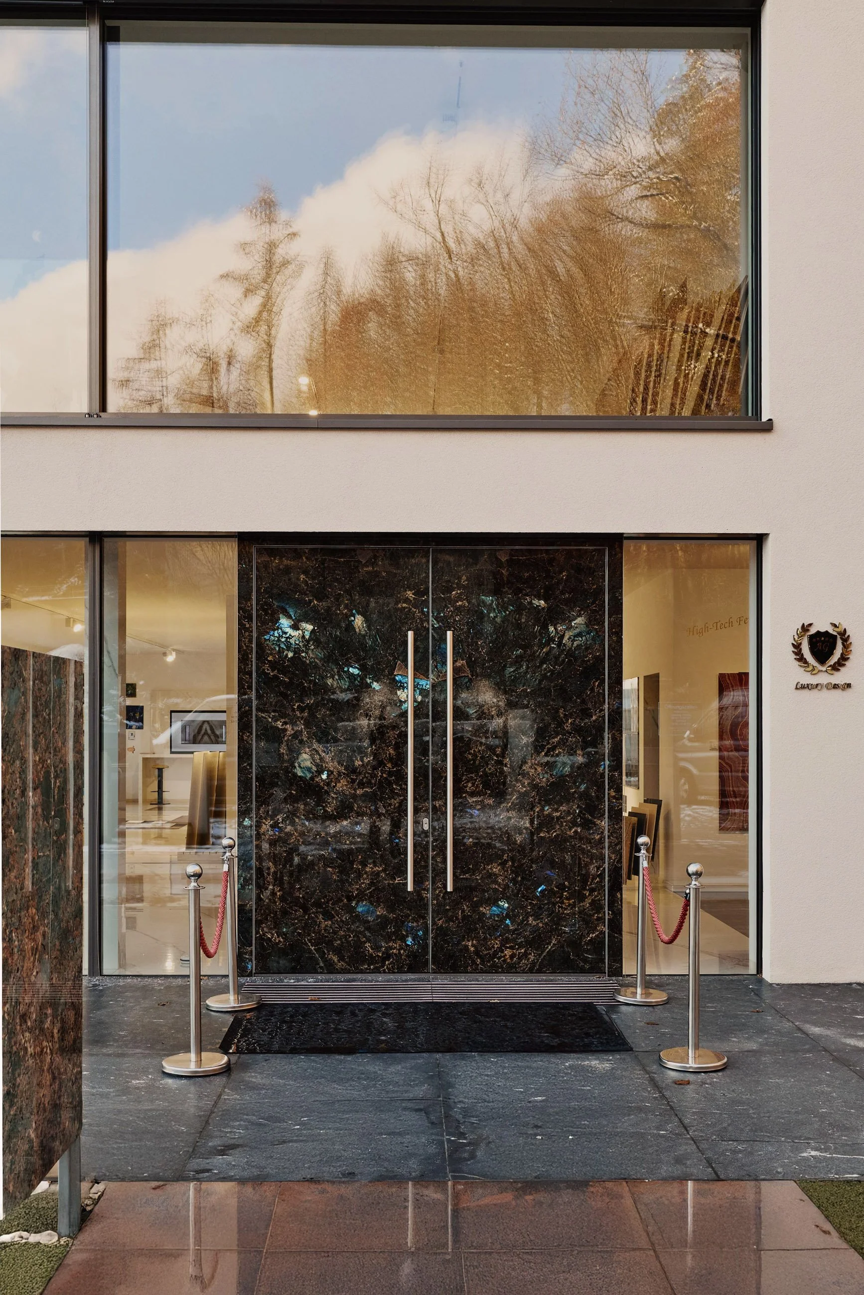 Eingang eines Gebäudes mit schwarzen Marmortüren, flankiert von Glasfenstern, und einer schwarzen Begrenzung am Boden. Das Gebäude hat ein modernes, elegantes Erscheinungsbild mit weißen Wänden und einem kleinen Schild rechts neben dem Eingang.