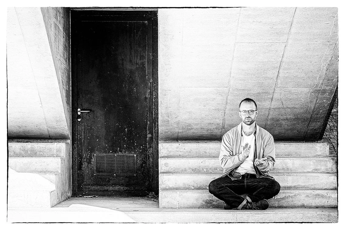 A man with glasses and a beard sitting cross-legged on steps next to a dark door, making a hand gesture.
