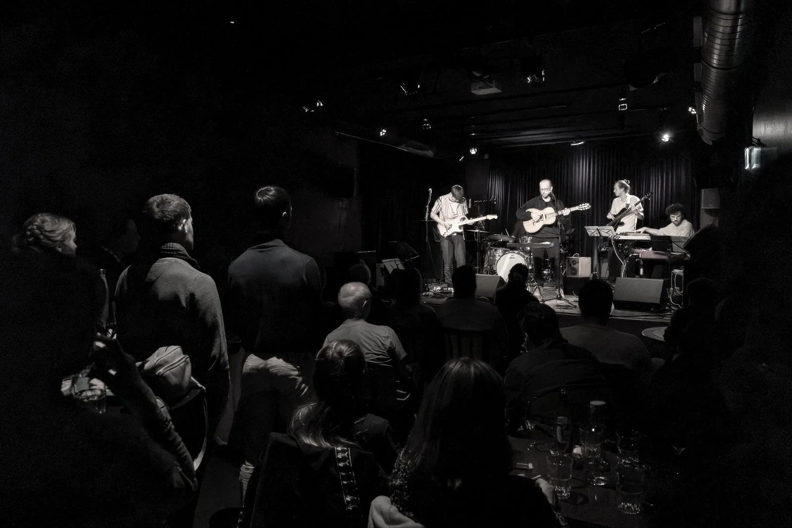 Black and white photo of a live music performance on a small stage with five musicians playing guitars, keyboard, and drums, audience watching in a dimly lit venue.