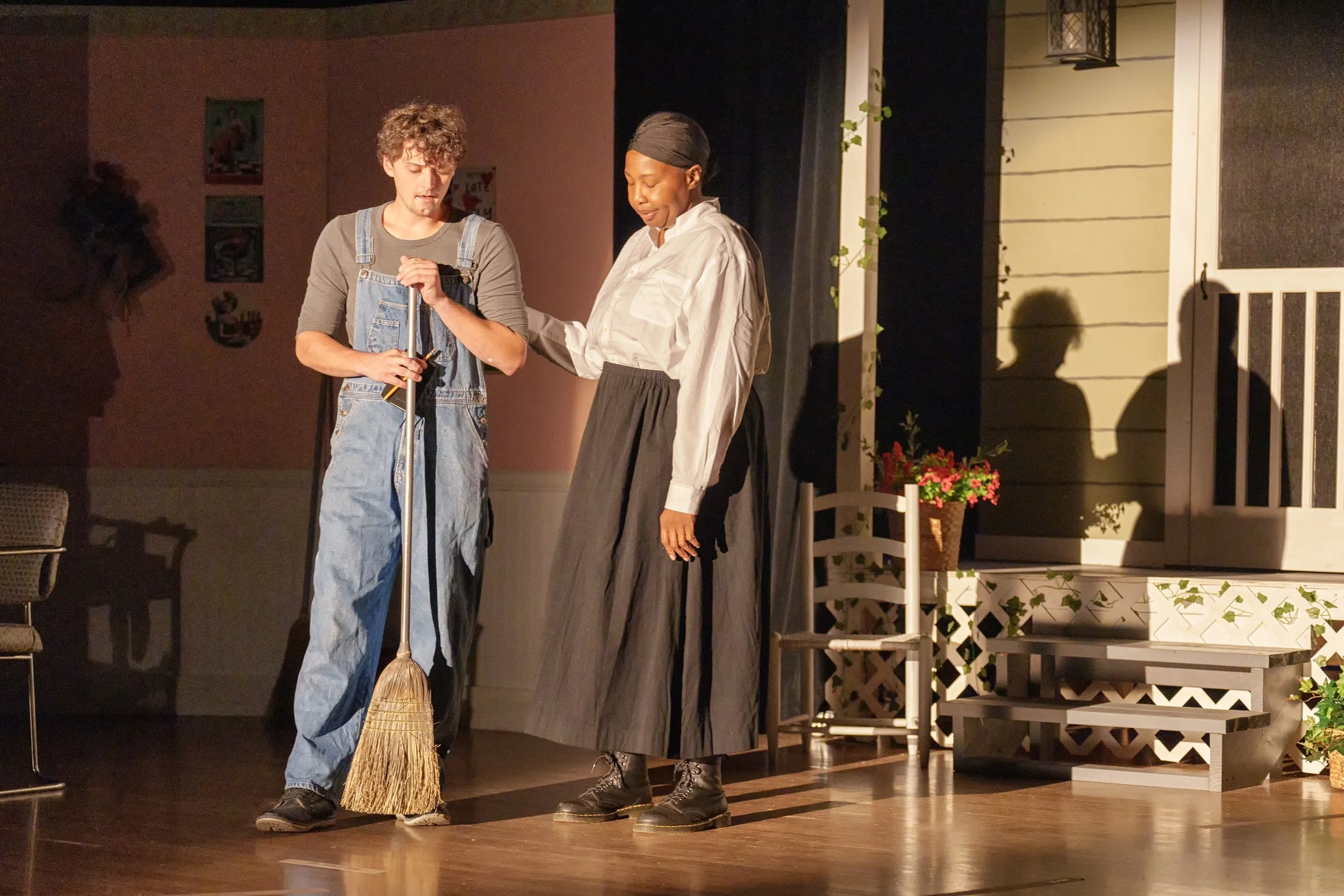 Two actors on stage, one with a broom and the other with a hand on the broom holder's shoulder, in a theatrical scene with a porch and plants in the background.