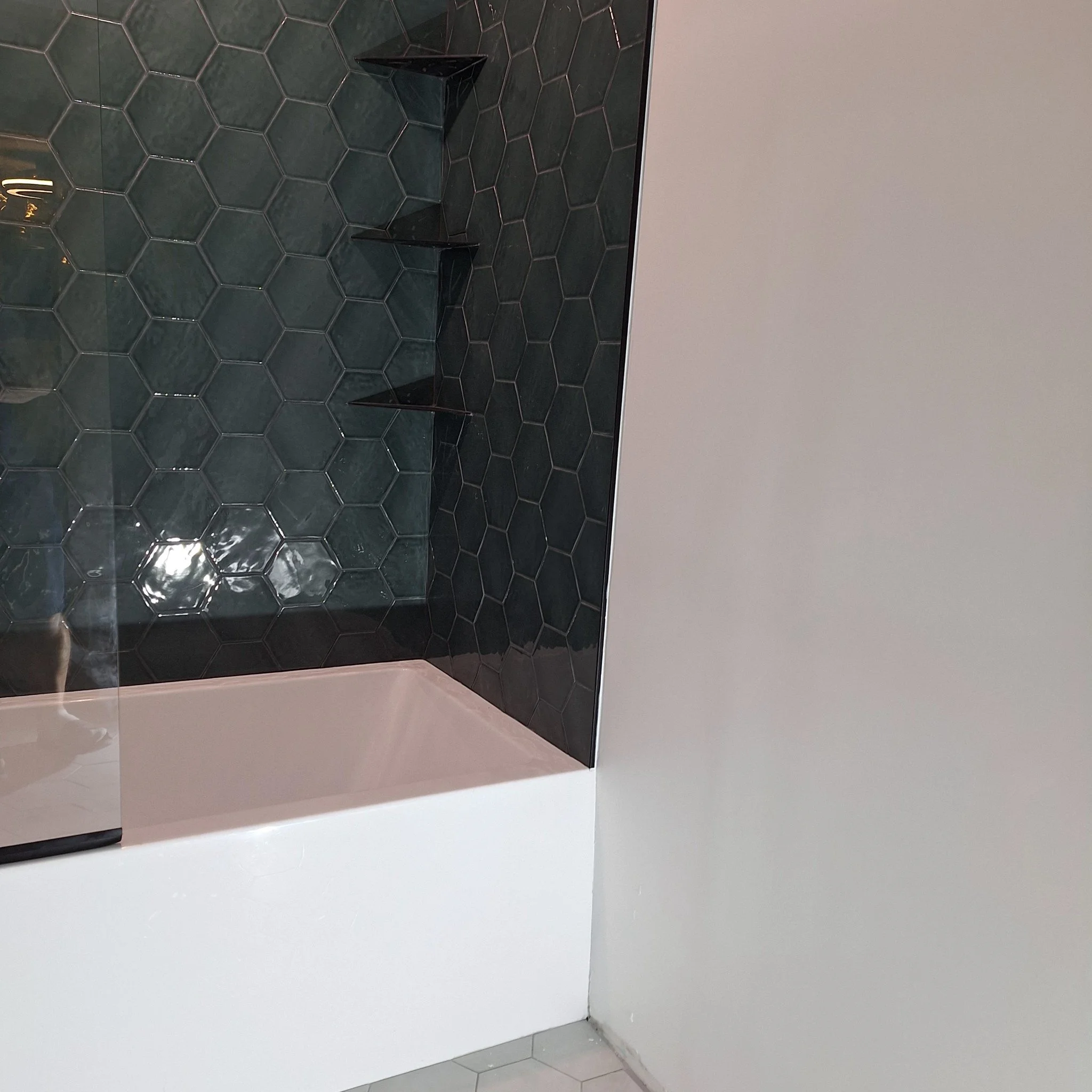 A modern bathroom shower area with black hexagonal tiles on the wall and a white bathtub. There are black corner shelves inside the shower.